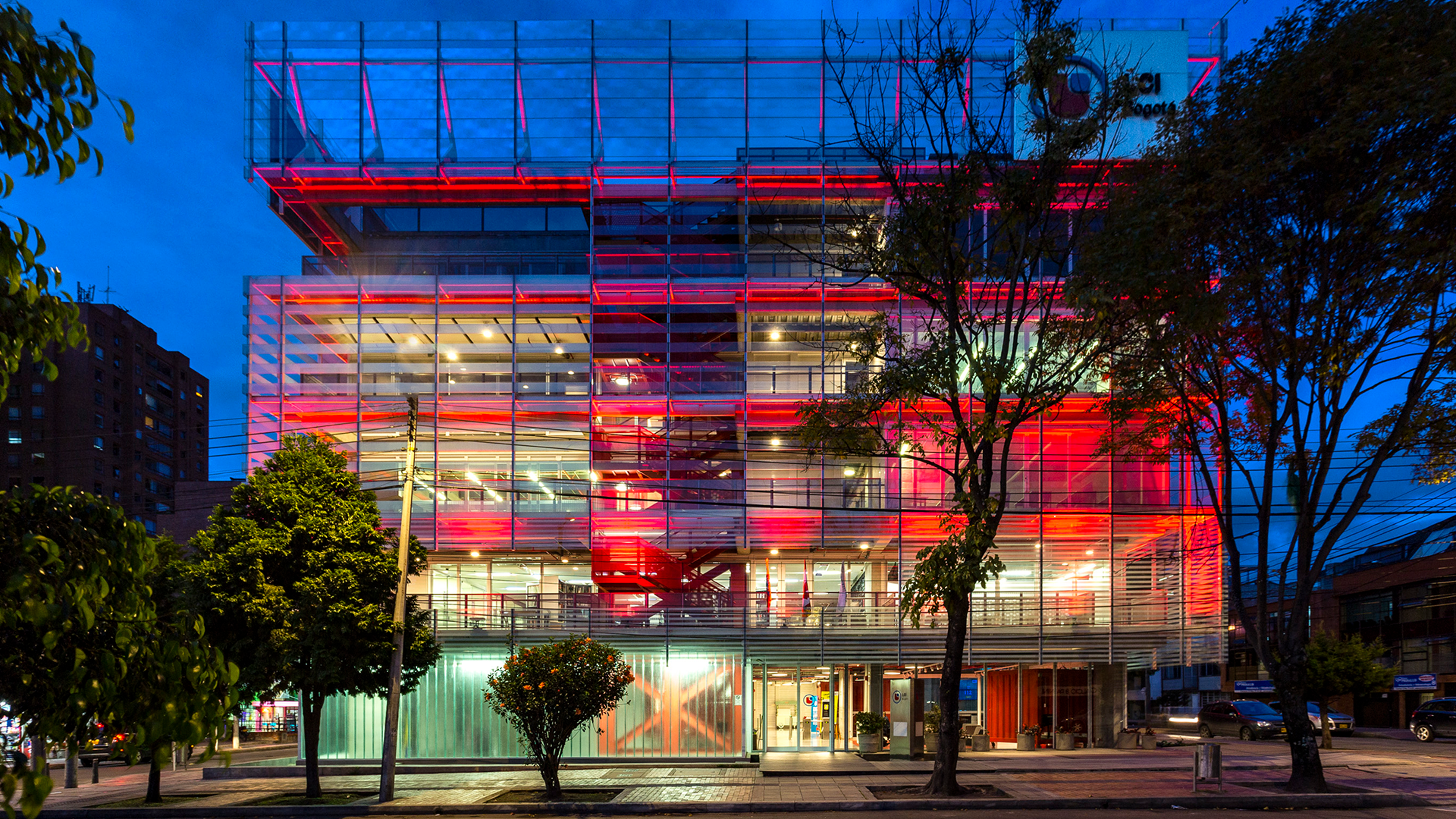 A contemporary building bathed in vibrant red lighting stands out against the evening sky, its glass facade reflecting the ambient city light.