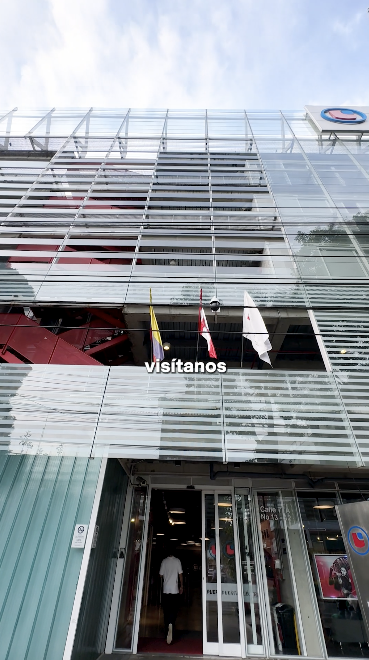 A modern building with glass and metal facades, featuring three flags hanging from the exterior and a person walking through the entrance.