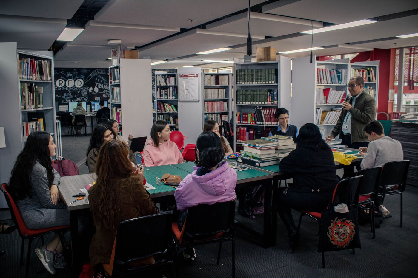 A group of students engaged in a study session at a library, with a person speaking at the head of the table.