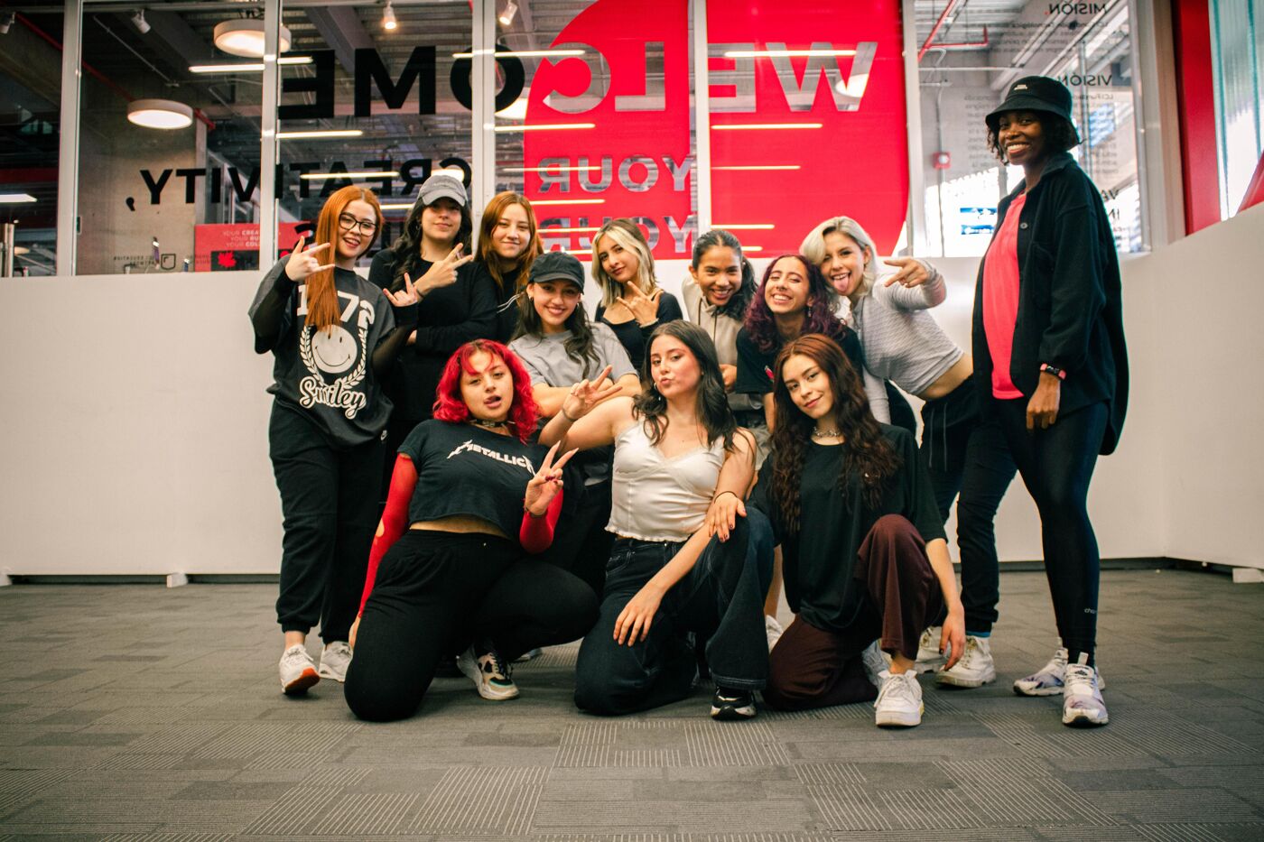 A group of approximately 12 young women are posing for a photo in front of a red and white sign. Most of them are dressed casually, and some are wearing hats. They appear to be in a dance studio or similar space.