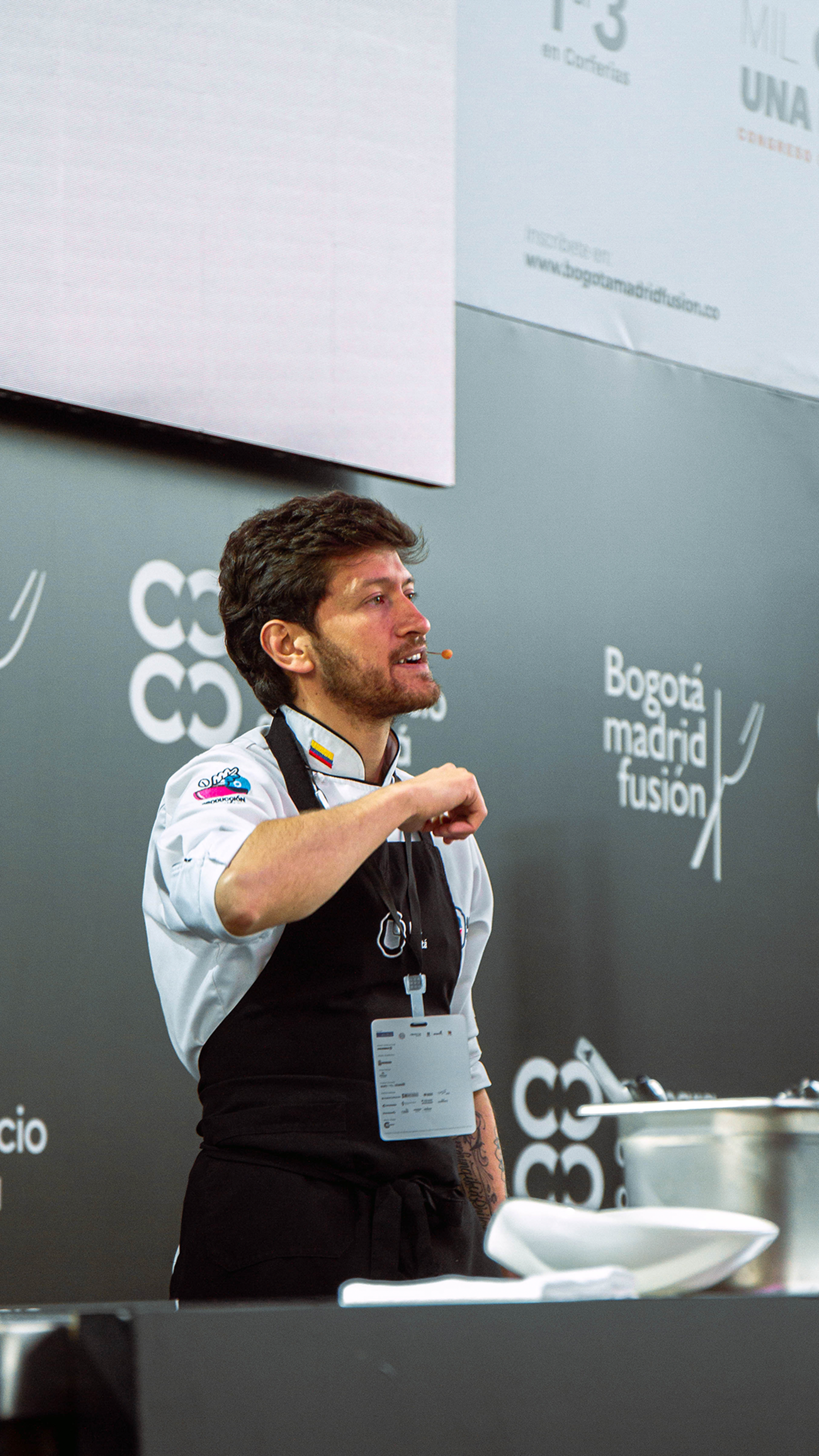 A chef in professional attire engages an audience during a culinary demonstration, with a microphone headset, against a simple backdrop.