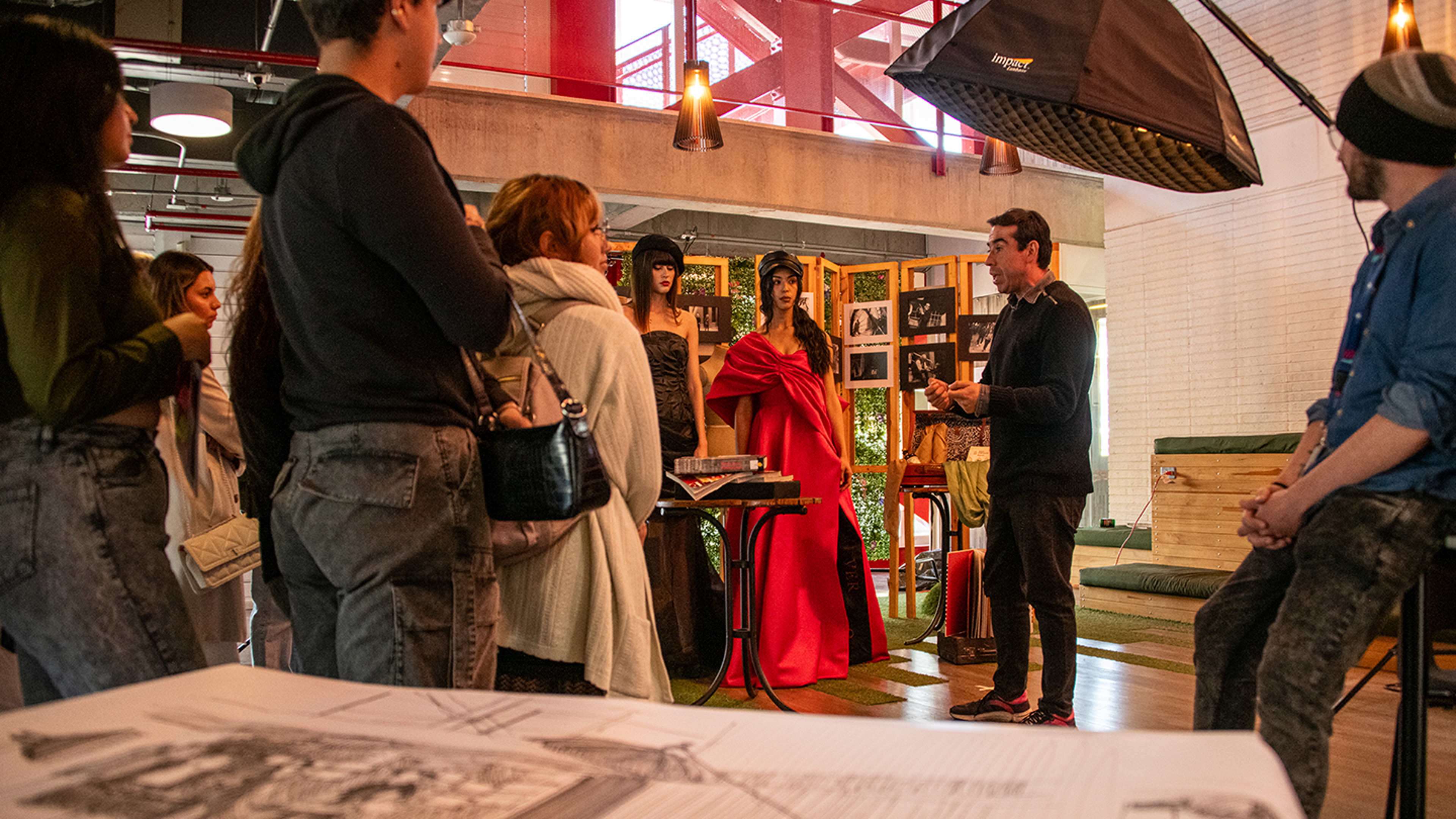 A presenter engages an audience at an art and fashion exhibition, with two models in elegant gowns central to the display.