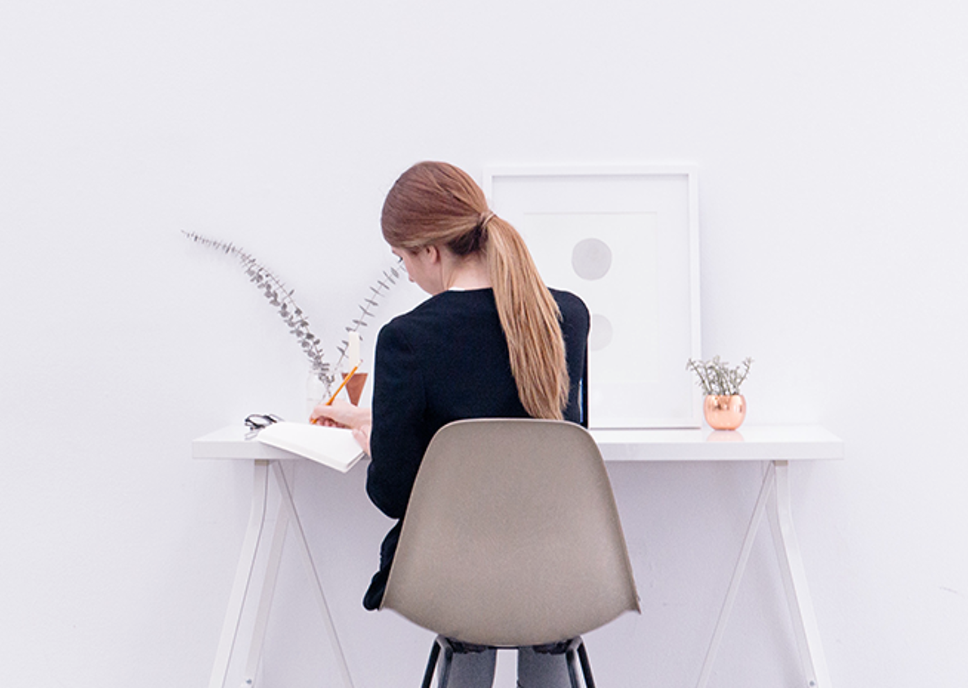 A woman with a ponytail is seated writing in a book at a minimalist white desk with simple decorations.