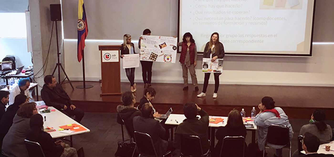 A group of people are gathered in a room, some presenting projects on stage while others are seated at tables, possibly engaged in a workshop or conference.