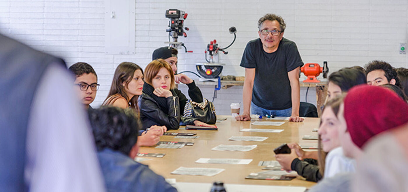 A focused group of students seated at a long table attentively listening to a standing lecturer in a casual classroom setting.