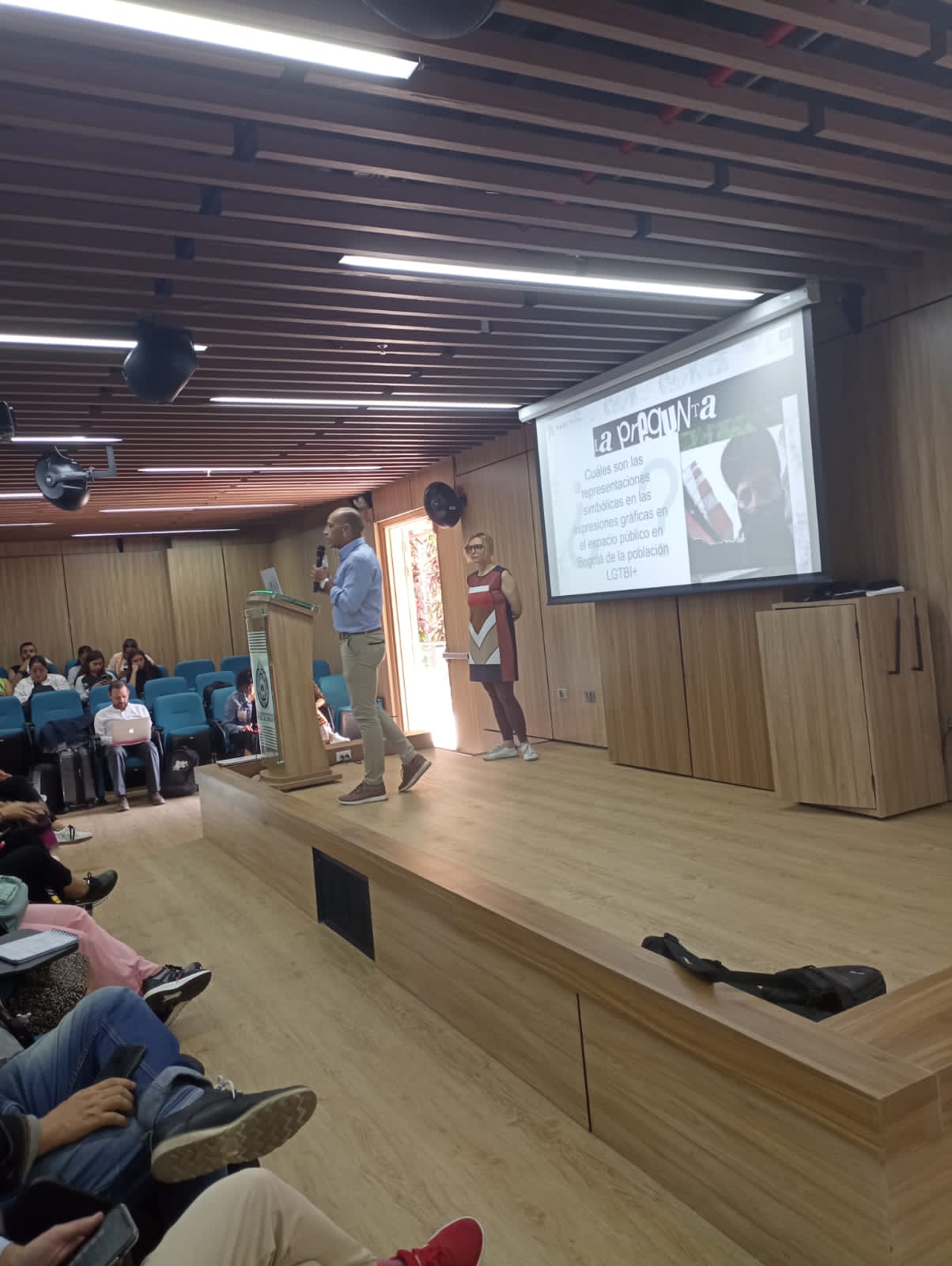 Dos presentadores, un hombre y una mujer, están junto a una pantalla grande que muestra una diapositiva en una sala de conferencias con asistentes.