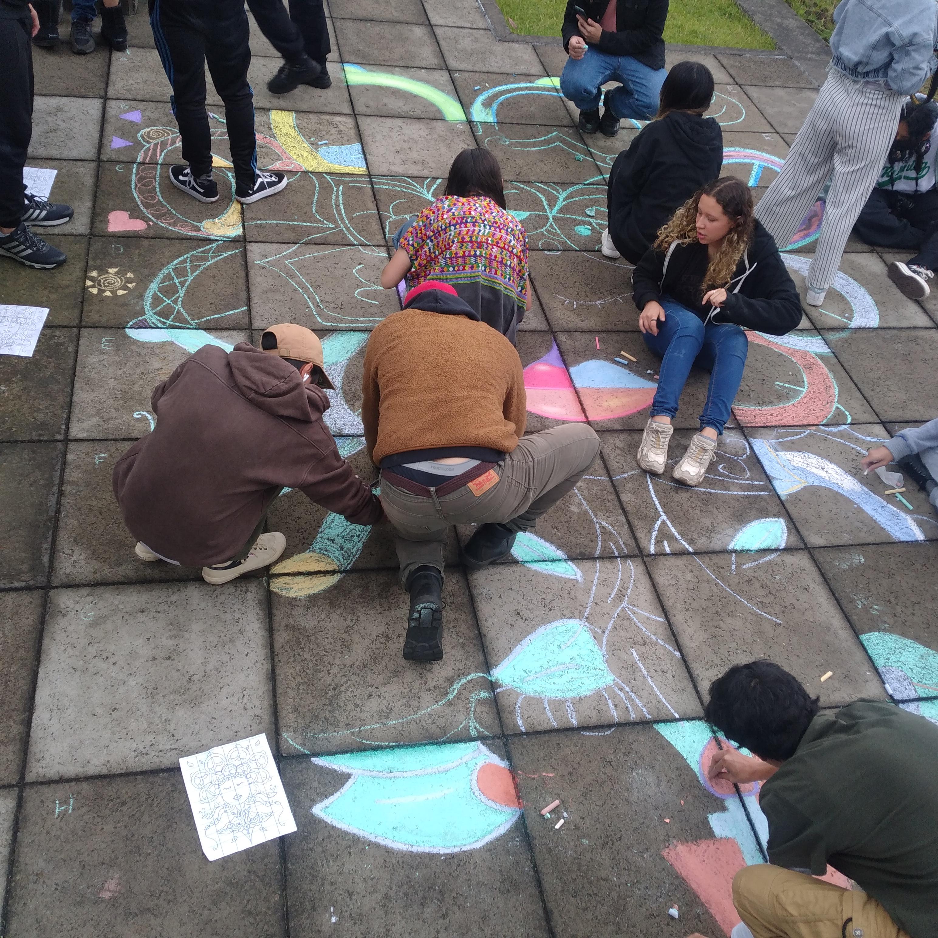 Personas dibujando un gran mural de tiza colorido en el pavimento, rodeado de espectadores.