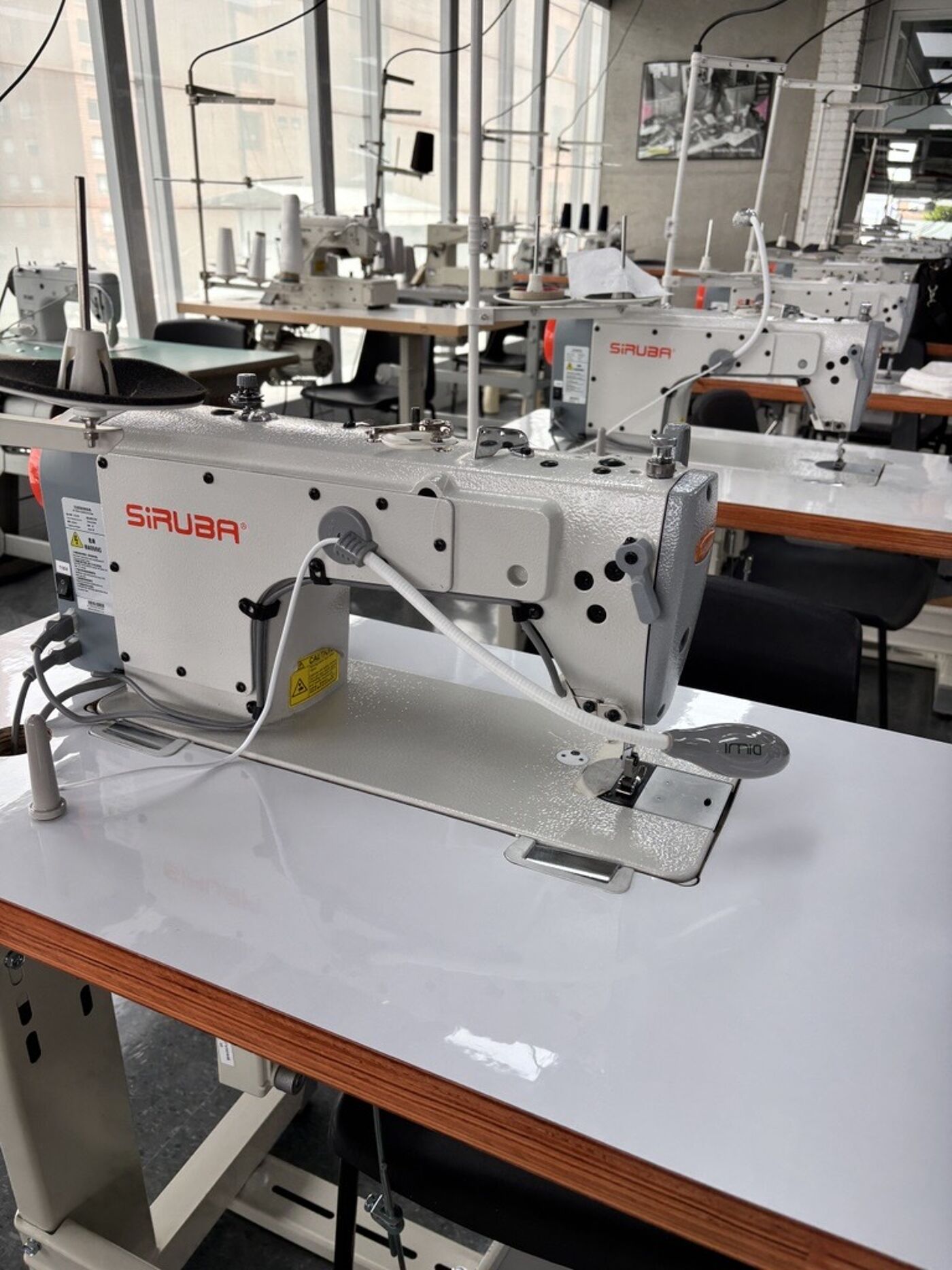 A Siruba industrial sewing machine is in the foreground, with other sewing machines and tables in a professional workshop in the background.