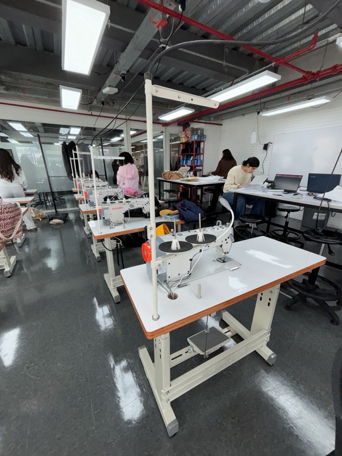 A sewing classroom featuring multiple industrial sewing machines and students actively engaged in their projects, creating a productive learning environment.