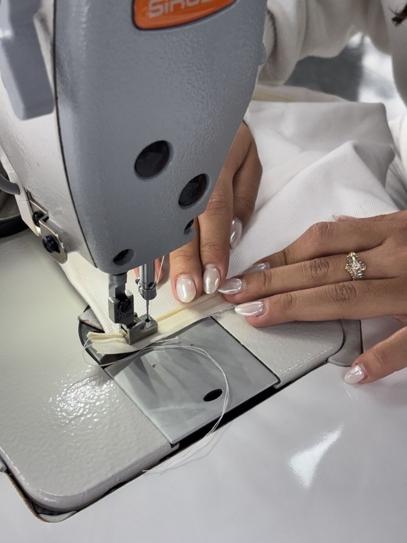 A person with well-manicured nails is using a sewing machine to attach a zipper to a cream-colored fabric. A diamond ring is visible on their finger.