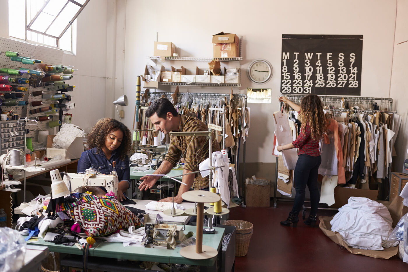 Three people are working in a sewing workshop, with sewing machines, fabrics, and clothes hanging on shelves. Two men and one woman are busy with their tasks.