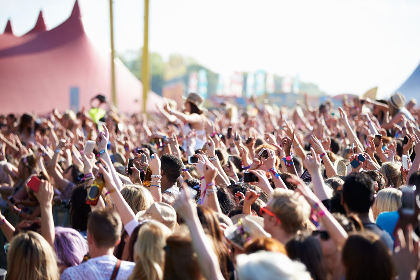 A lively crowd enjoys an outdoor concert under a clear sky, with tents and stages in the background, creating a vibrant festival atmosphere.