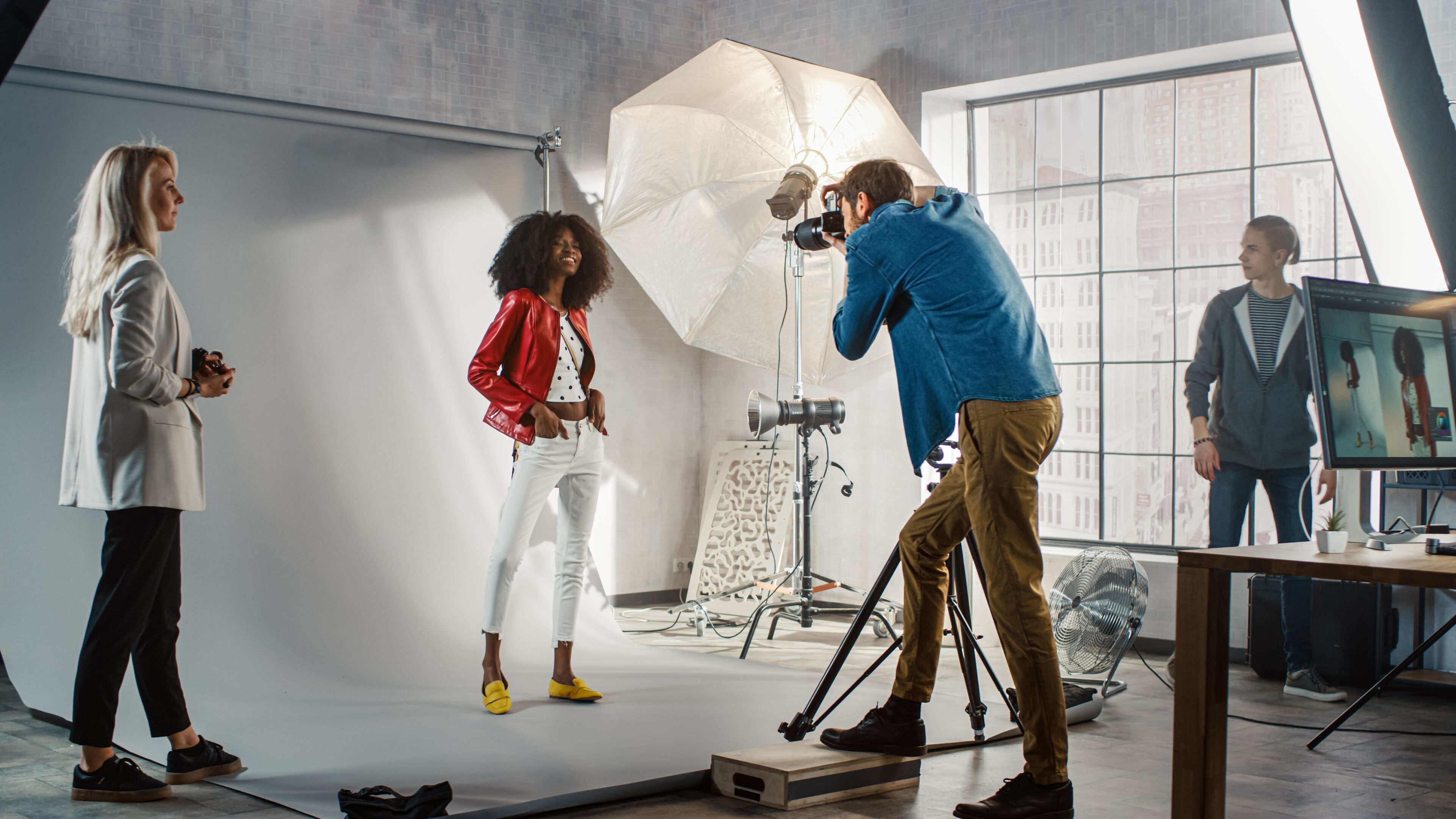 Un fotógrafo toma una foto de una modelo con chaqueta roja y pantalón blanco, con asistentes que manejan el equipo en un estudio, con la ciudad de fondo.