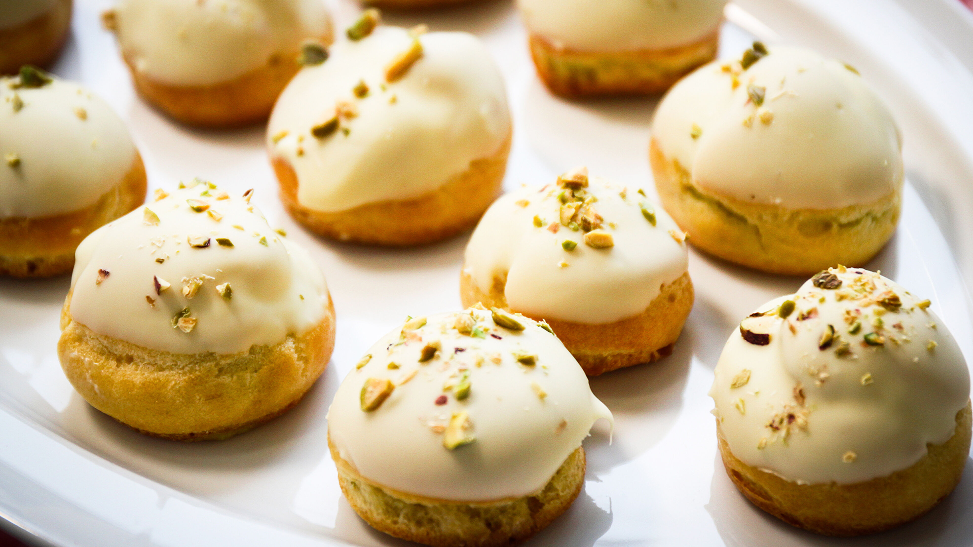 Elegantes pasteles con glaseado blanco y pistachos, presentados en una bandeja blanca.