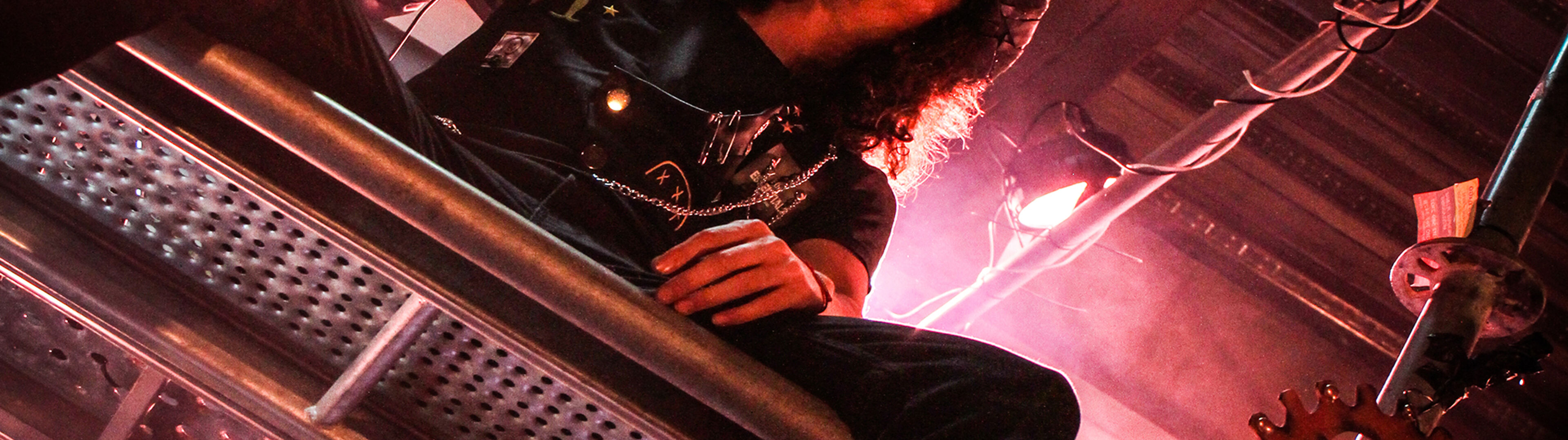Músico con cabello rizado en un escenario iluminado por luces cálidas, rodeado de un entorno de temática industrial.