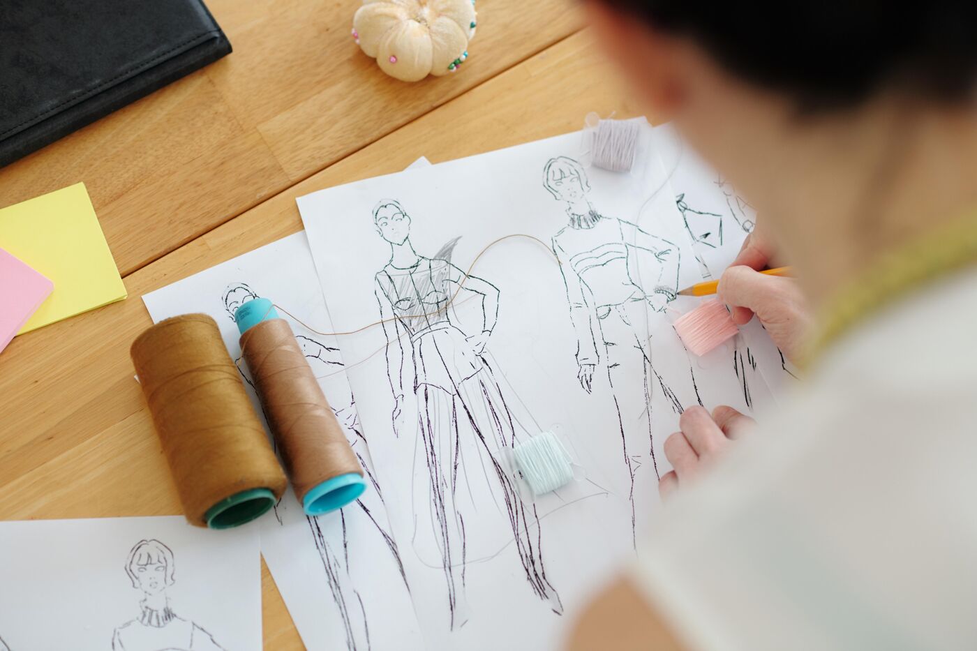 The image shows a person sketching fashion designs on paper. There are spools of thread and other design elements on the table, suggesting a creative workspace.