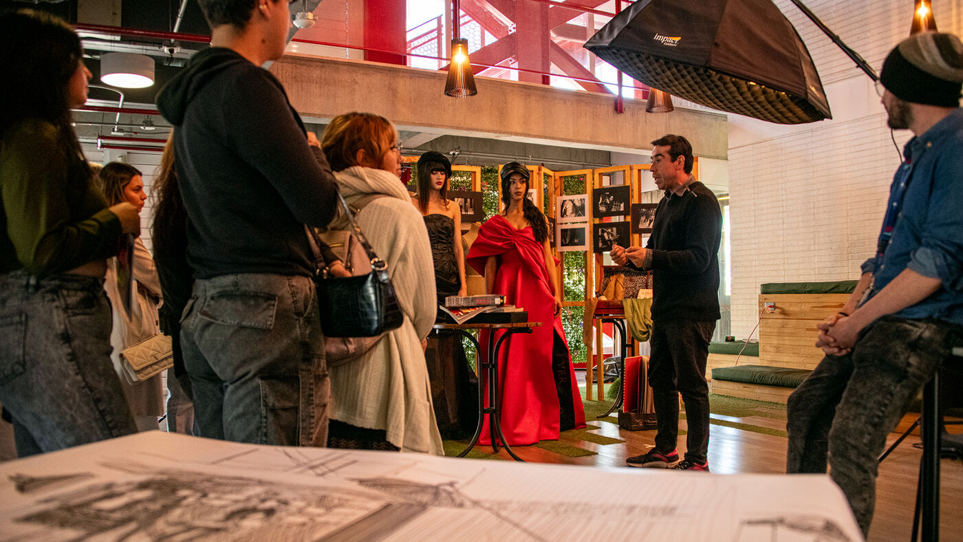 People gather at an indoor art exhibit opening, viewing photographs and listening to a speaker.