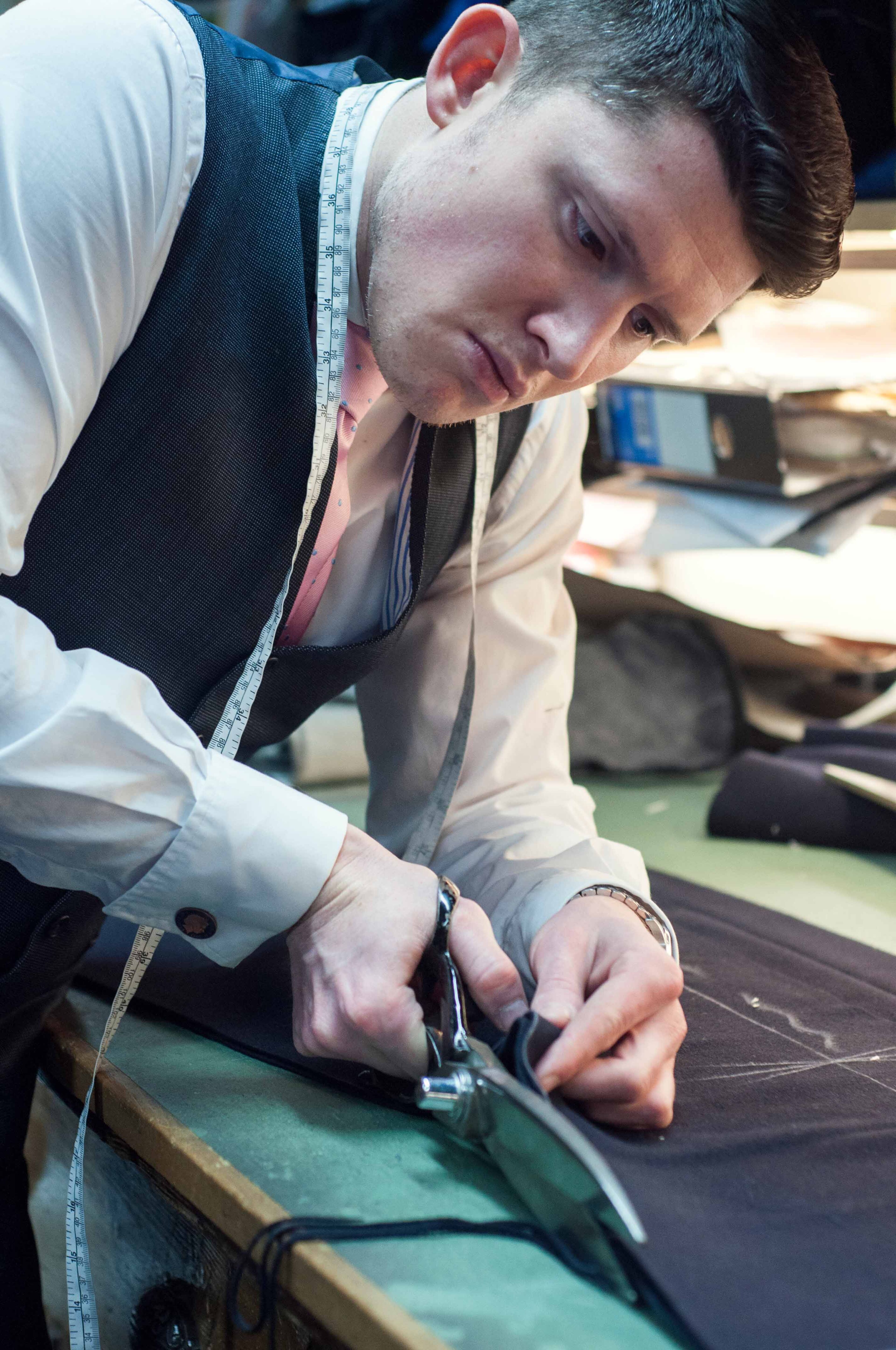 Un sastre enfocado recorta tela con tijeras en un banco de trabajo, con una cinta métrica alrededor de su cuello.