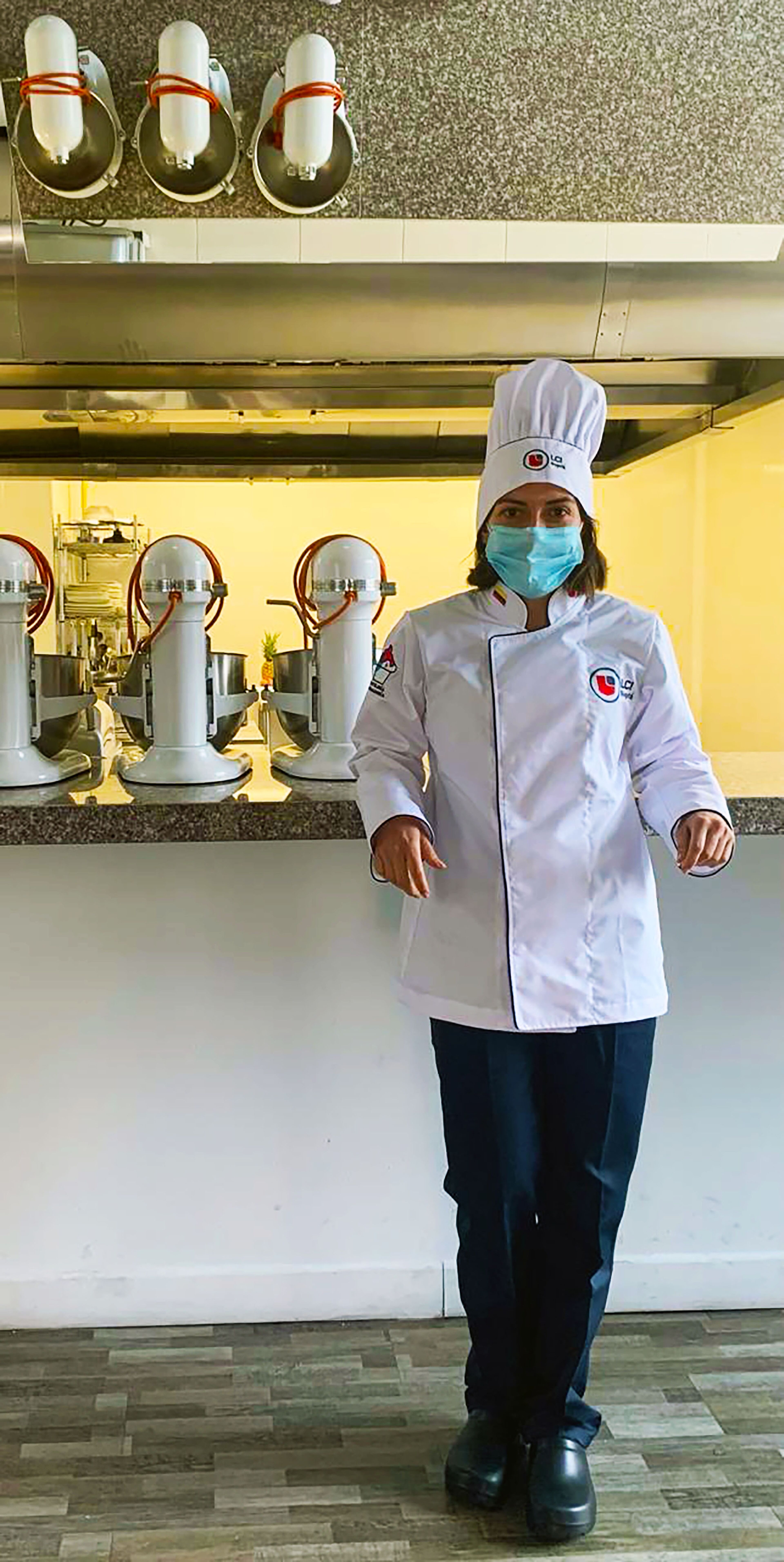 Un(a) chef profesional en uniforme blanco y gorro se encuentra en una cocina comercial, llevando mascarilla por seguridad sanitaria.
