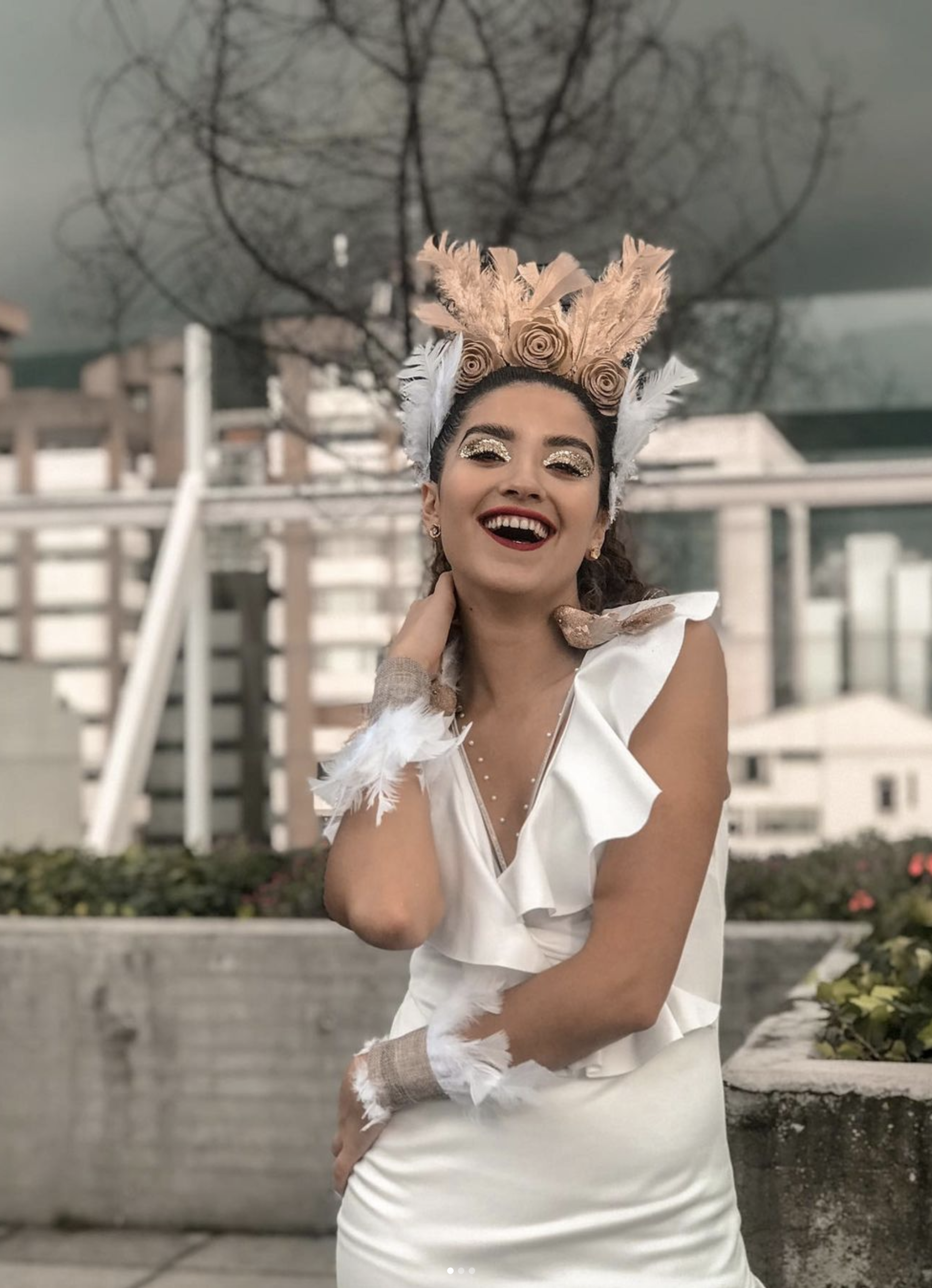 Una mujer con una sonrisa radiante, vestida con un vestido blanco y un tocado creativo con plumas y rosas, irradia alegría contra un telón de fondo urbano.