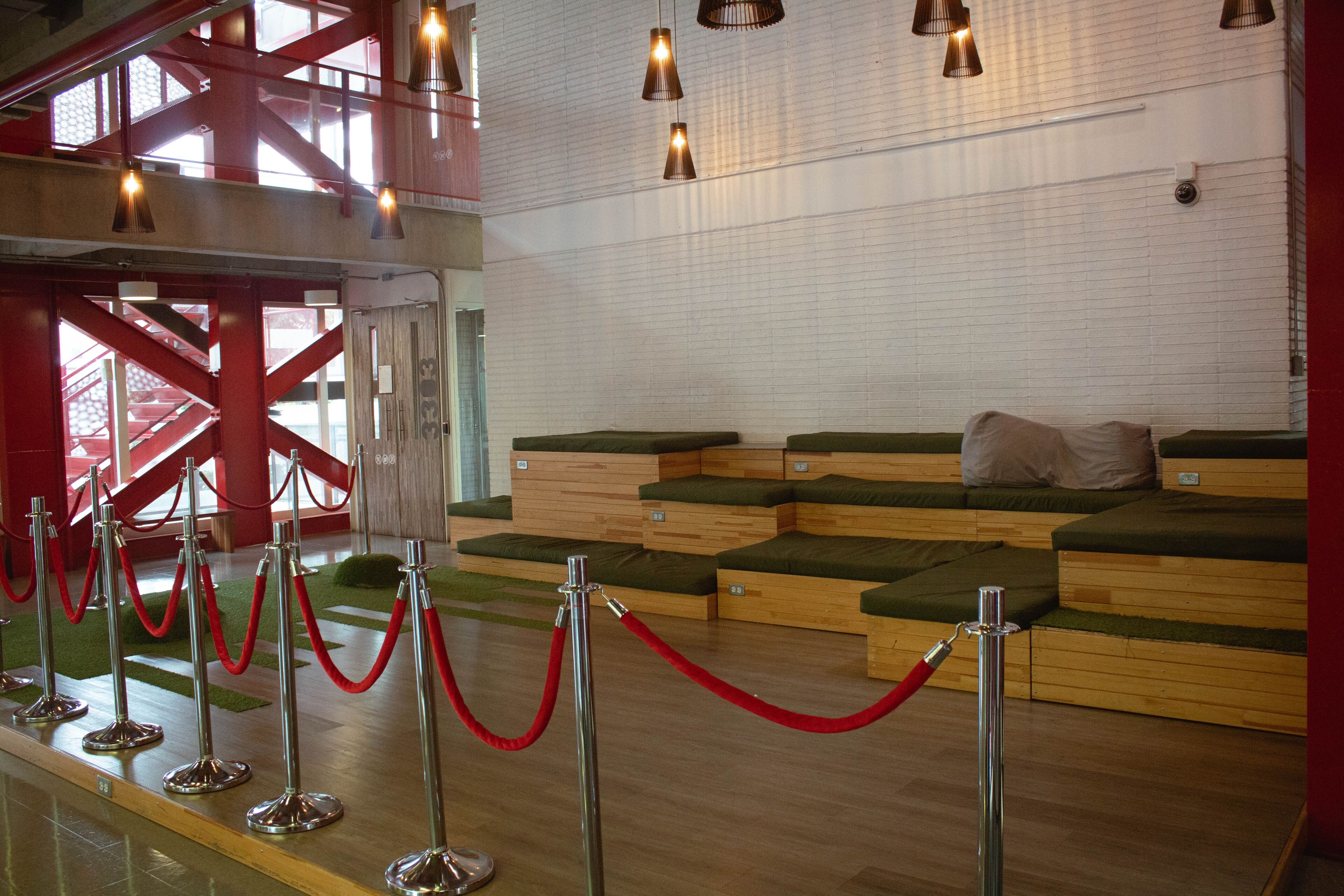 Espacio interior con estilo, con asientos de madera escalonados con cojines, delimitados por cuerdas de terciopelo y iluminación colgante industrial, junto a una escalera.