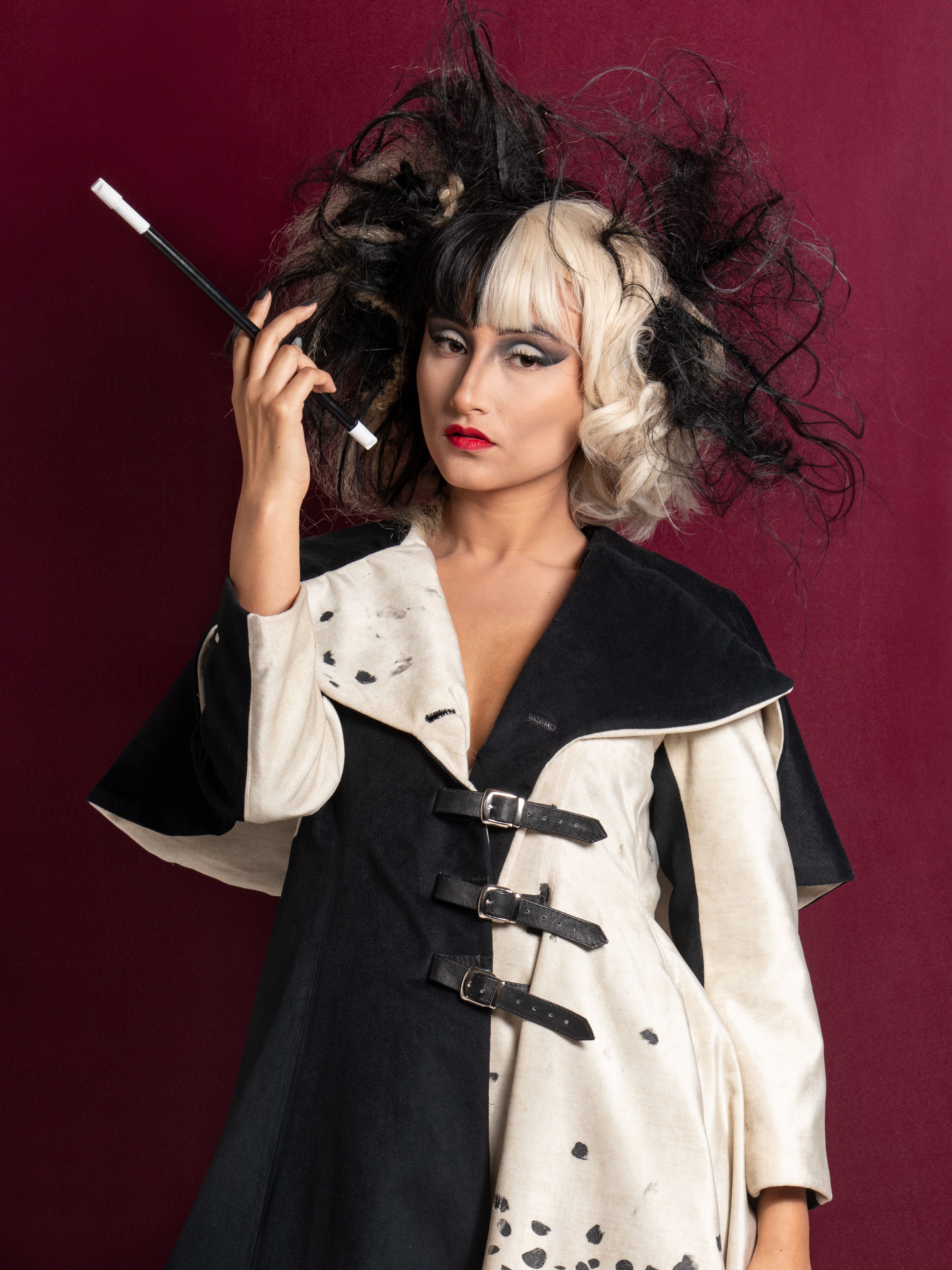 Persona posando dramáticamente, evocando la icónica apariencia de Cruella de Vil con un peinado en dos tonos, maquillaje intenso y abrigo moteado.