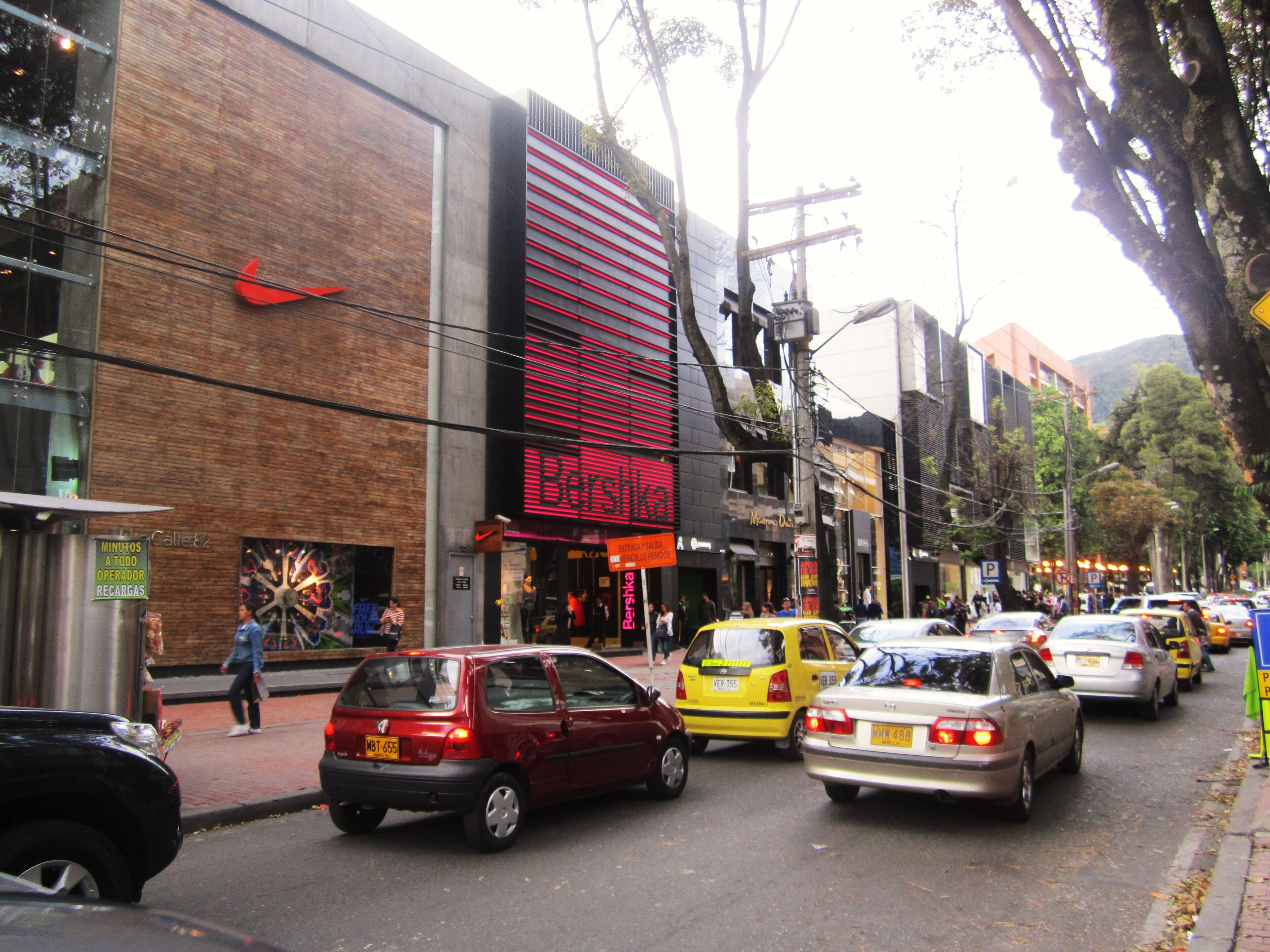 A bustling city street lined with retail stores, including a prominent sportswear outlet and a fashion retailer. Cars and taxis navigate the traffic, with pedestrians walking along the sidewalk under the shade of street trees.