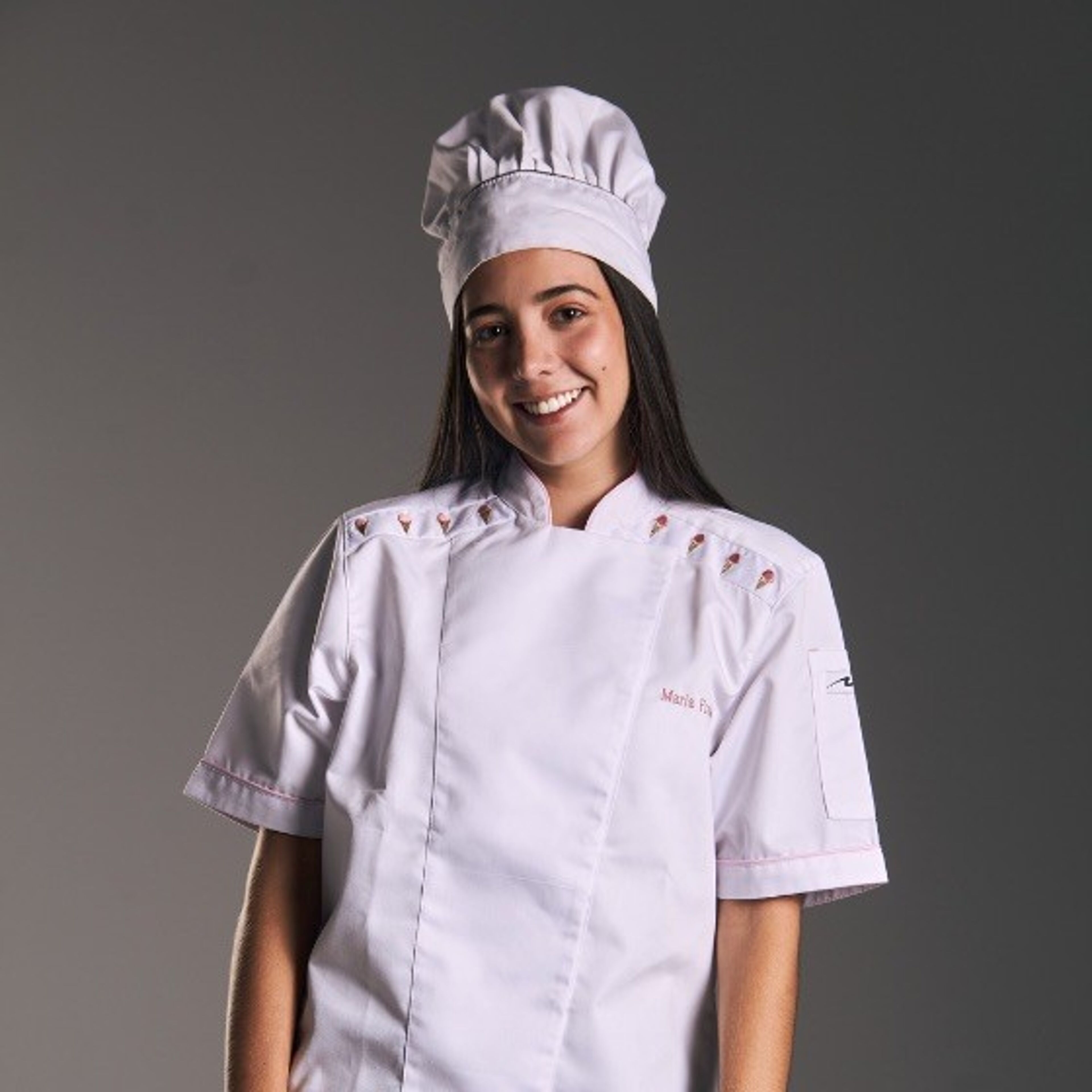 Una joven sonriente vestida con un uniforme blanco de chef profesional y un gorro, señalando su papel en las artes culinarias.