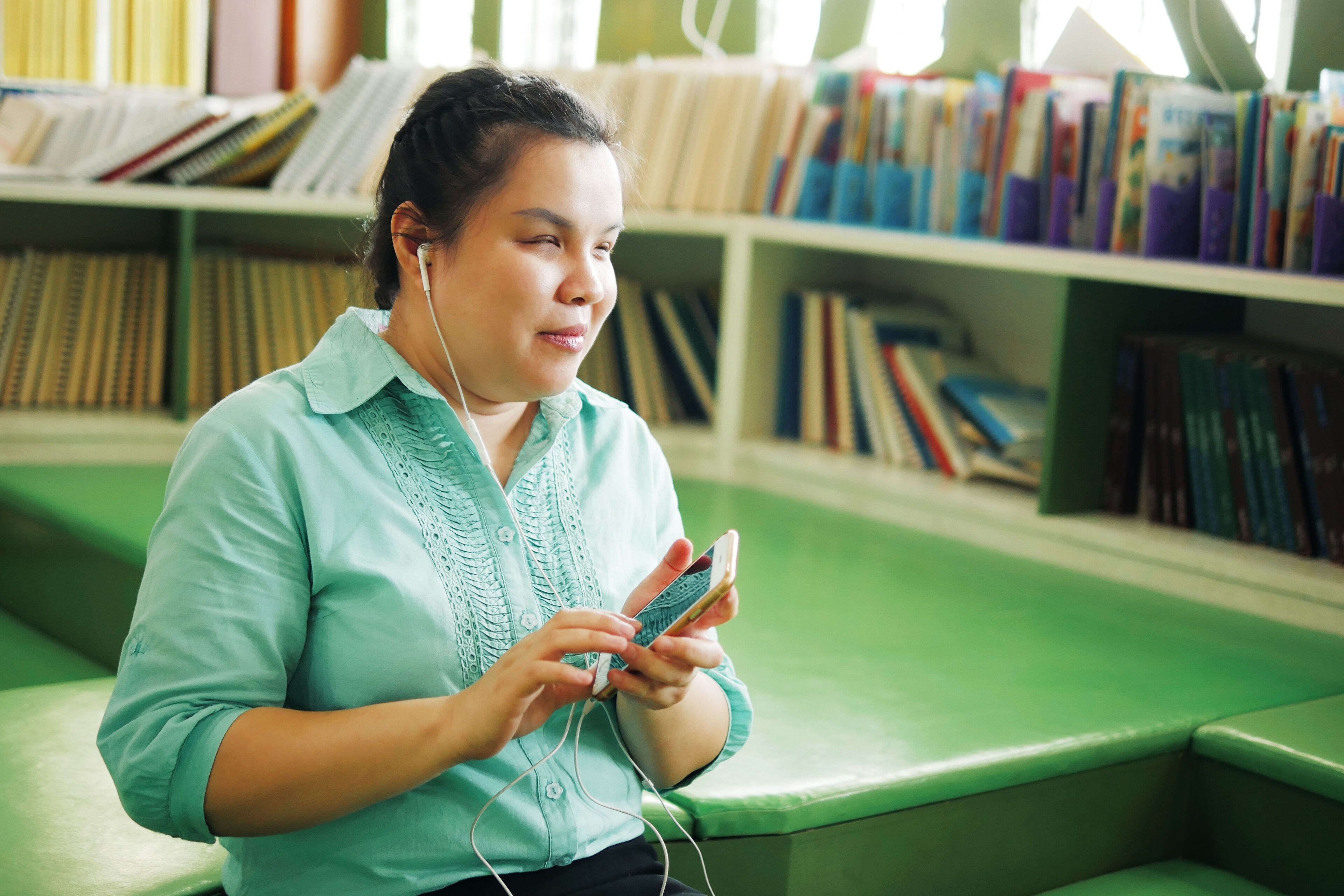 Portrait of Asian woman with blindness disability wearing earphones using smart phone with voice accessibility for persons with visual impairment disabilities in creative workplace library.