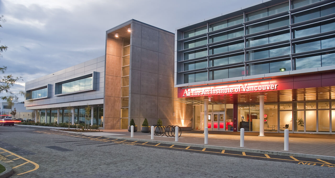 The modern exterior of The Art Institute of Vancouver at dusk, featuring sleek glass windows and concrete walls. A red neon sign marks the entrance, with warm lighting creating a welcoming atmosphere against the cloudy evening sky