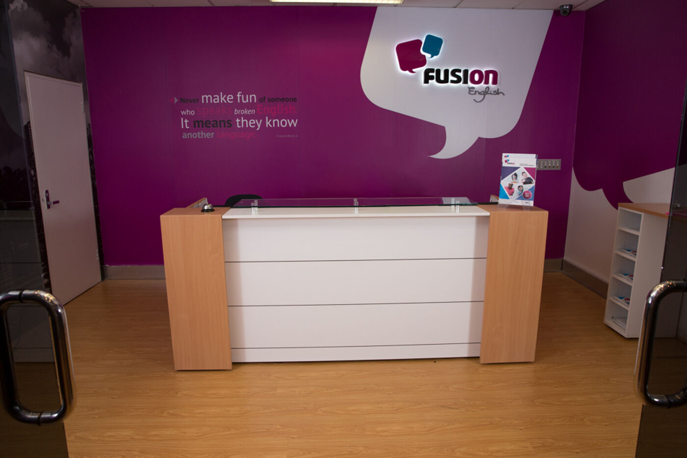 The reception area of Fusion English, featuring a modern white desk against a purple wall with motivational text promoting language learning. The branding and warm wood flooring create an inviting atmosphere for students.