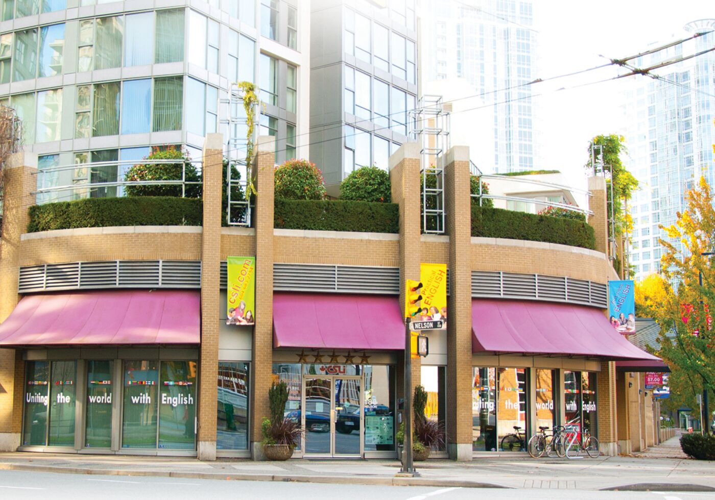 The vibrant exterior of a language school with purple awnings and banners promoting English programs. The modern building features large glass windows and a rooftop garden, creating a welcoming urban presence.