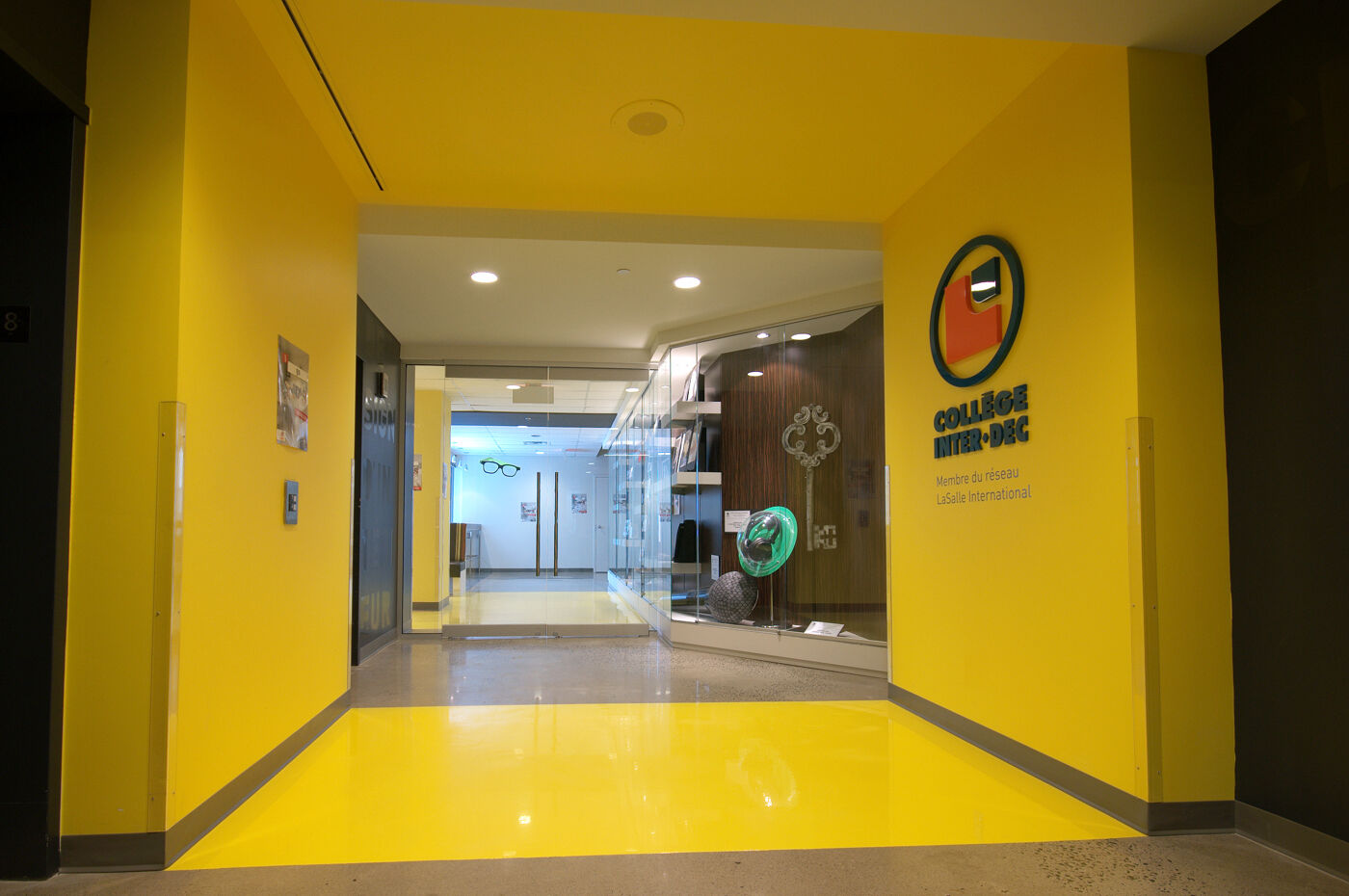 The vibrant yellow entrance hallway of Collège Inter-Dec, part of the LaSalle International network, features modern decor and glass display cases. The college logo is prominently displayed, adding to the lively and welcoming ambiance.