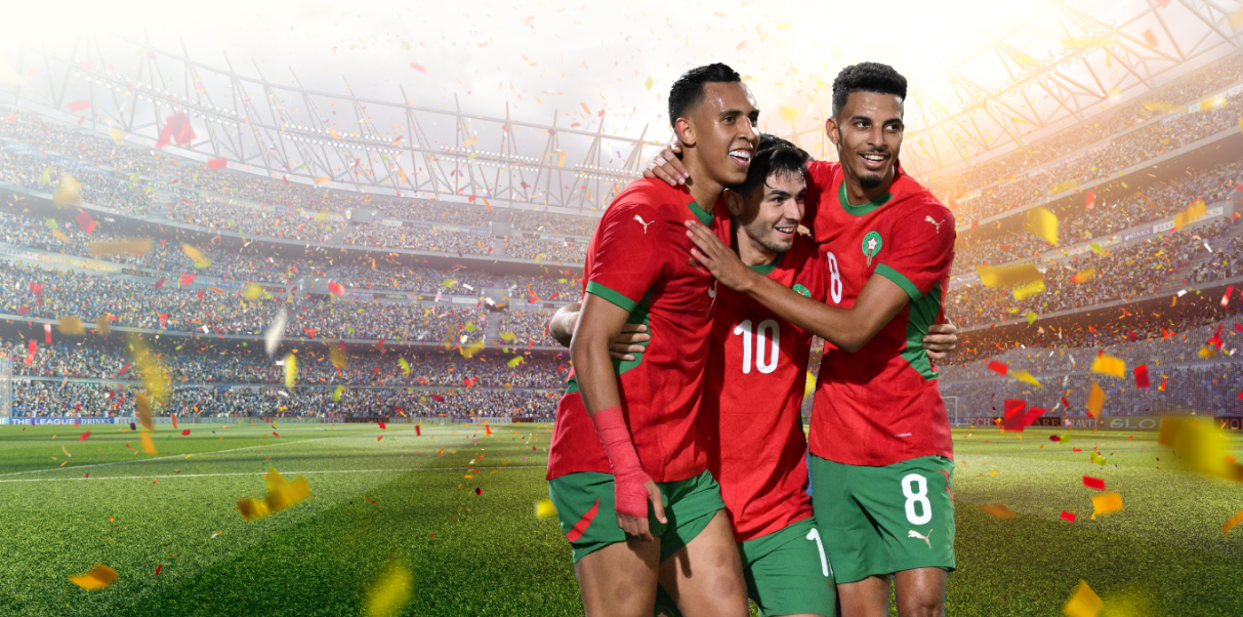 Three Moroccan soccer players in red and green uniforms embrace on a soccer field amidst celebratory confetti and a cheering stadium crowd.