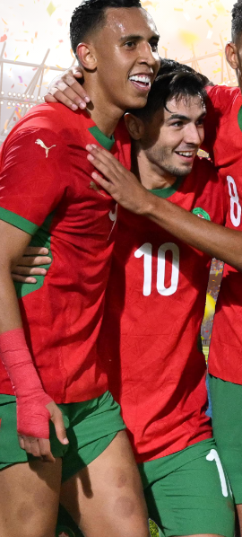 Three Moroccan soccer players in red and green uniforms embrace on a soccer field amidst celebratory confetti and a cheering stadium crowd.