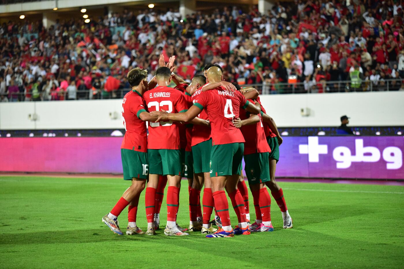 A group of soccer players in red and green uniforms celebrate a goal, huddled together on a green field in front of a large stadium crowd and a purple advertising board.