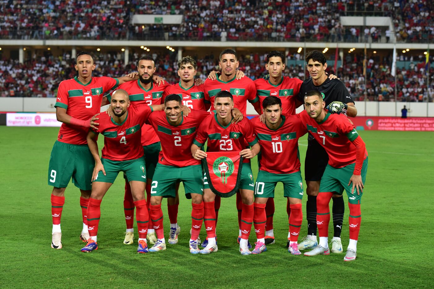 A soccer team, predominantly clad in red and green uniforms, stands shoulder-to-shoulder on a green field, their expressions serious as they pose for a photograph.