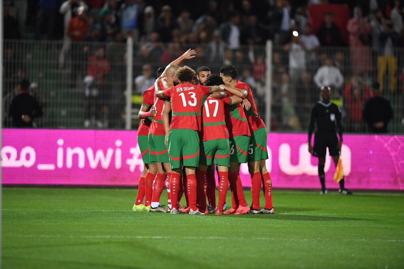 A group of soccer players in green and red uniforms huddle together on a field, celebrating a goal, with a referee and spectators visible in the background.