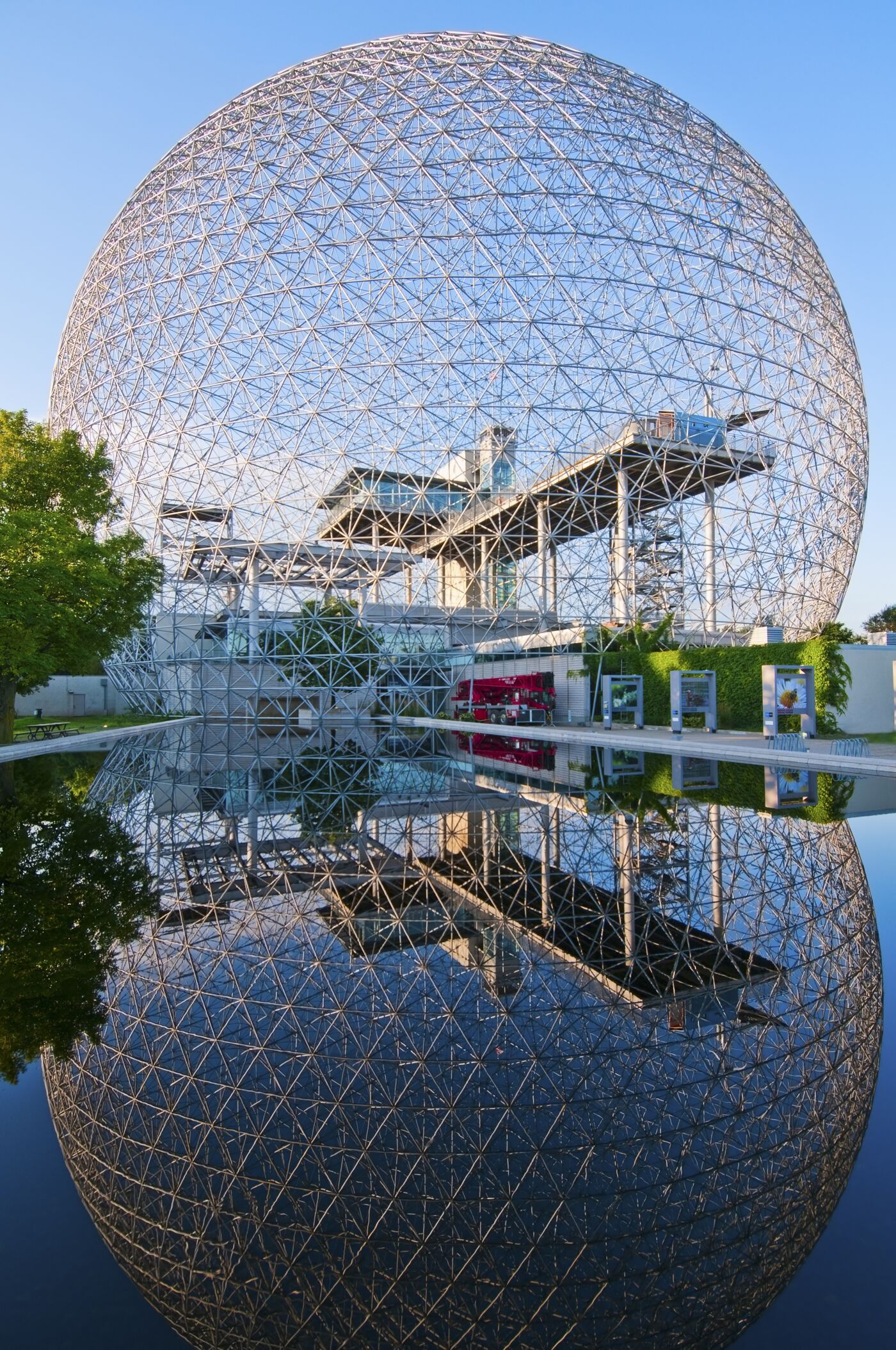 The intricate geodesic dome structure reflected on a calm water surface, set against a clear sky.