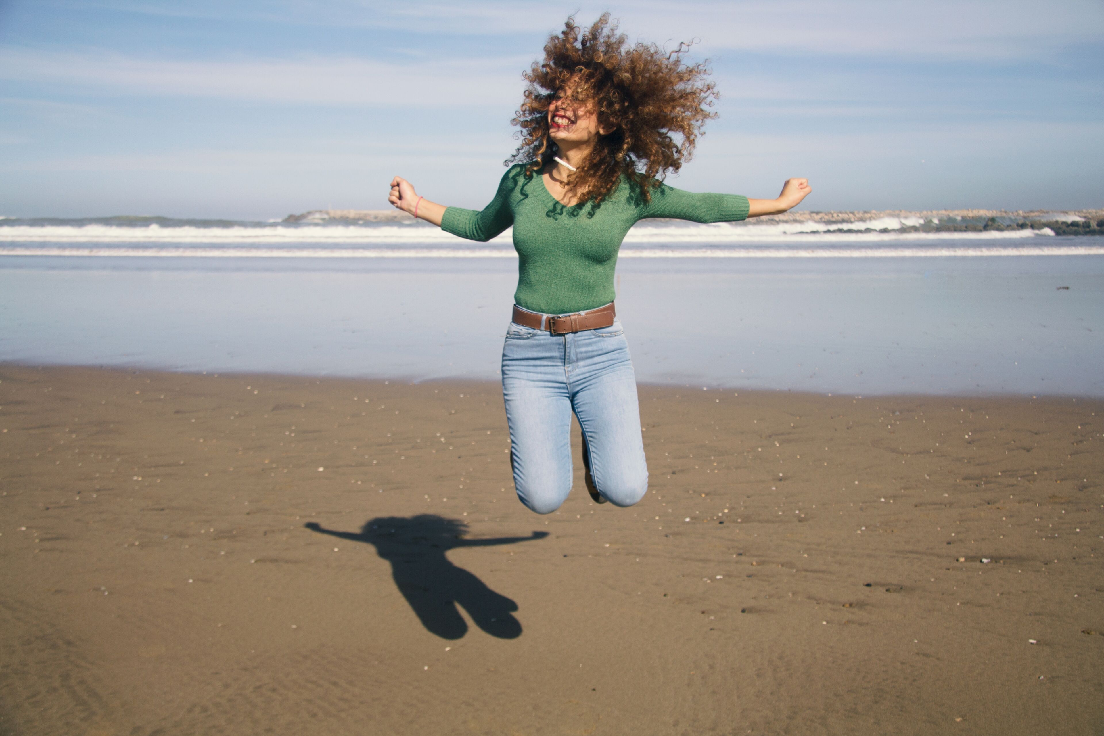 A person with curly hair in mid-jump, smiling broadly, against a beach backdrop with gentle waves.