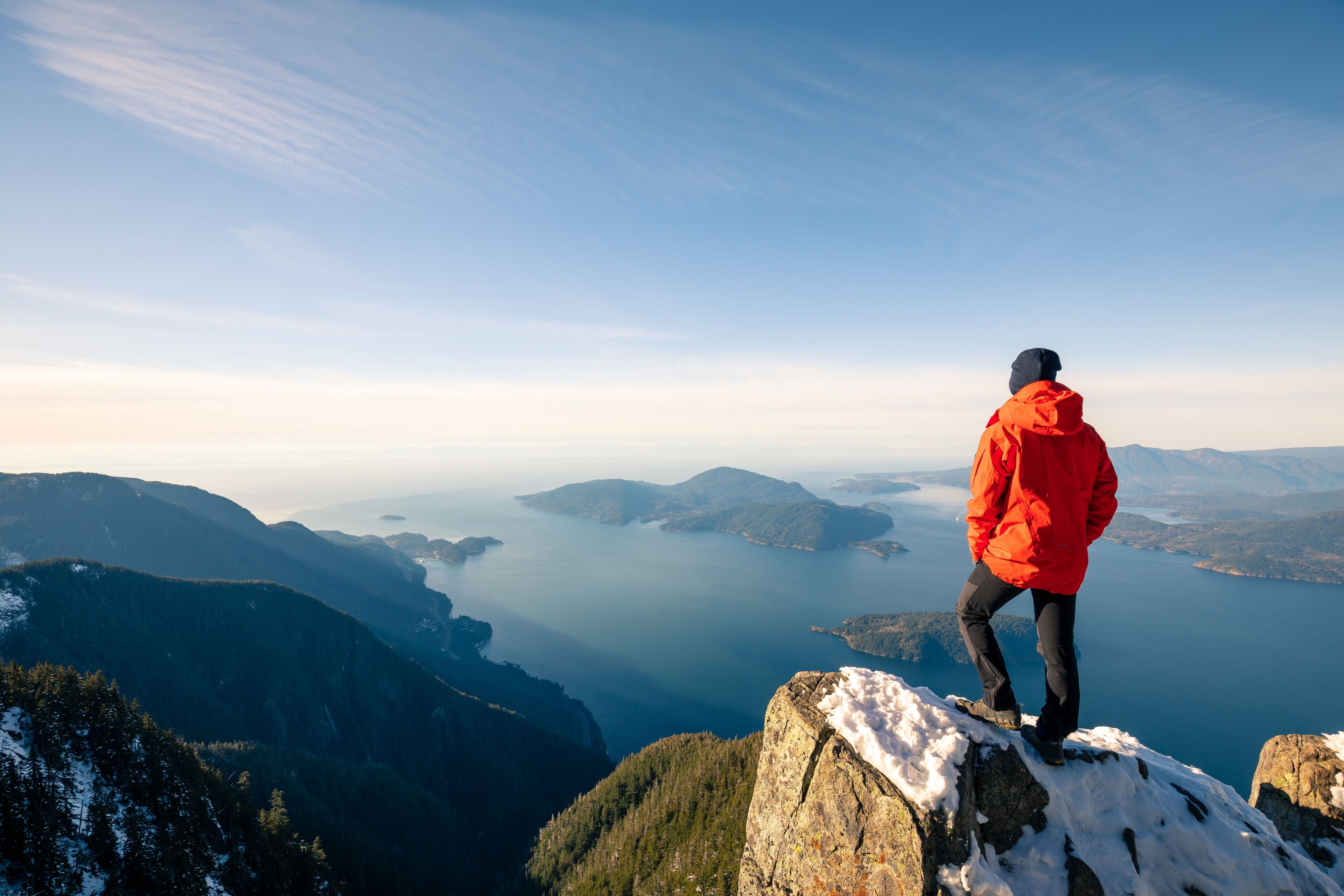 Man standing on mountain peak with snow overlooking ocean and islands in Vancouver, Canada.