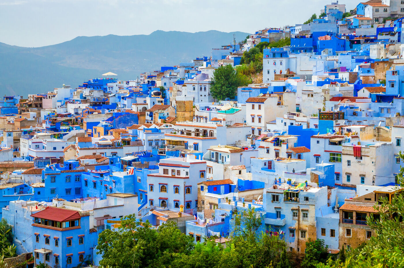 The image features a colorful city with blue buildings, located on a hillside, surrounded by mountains.