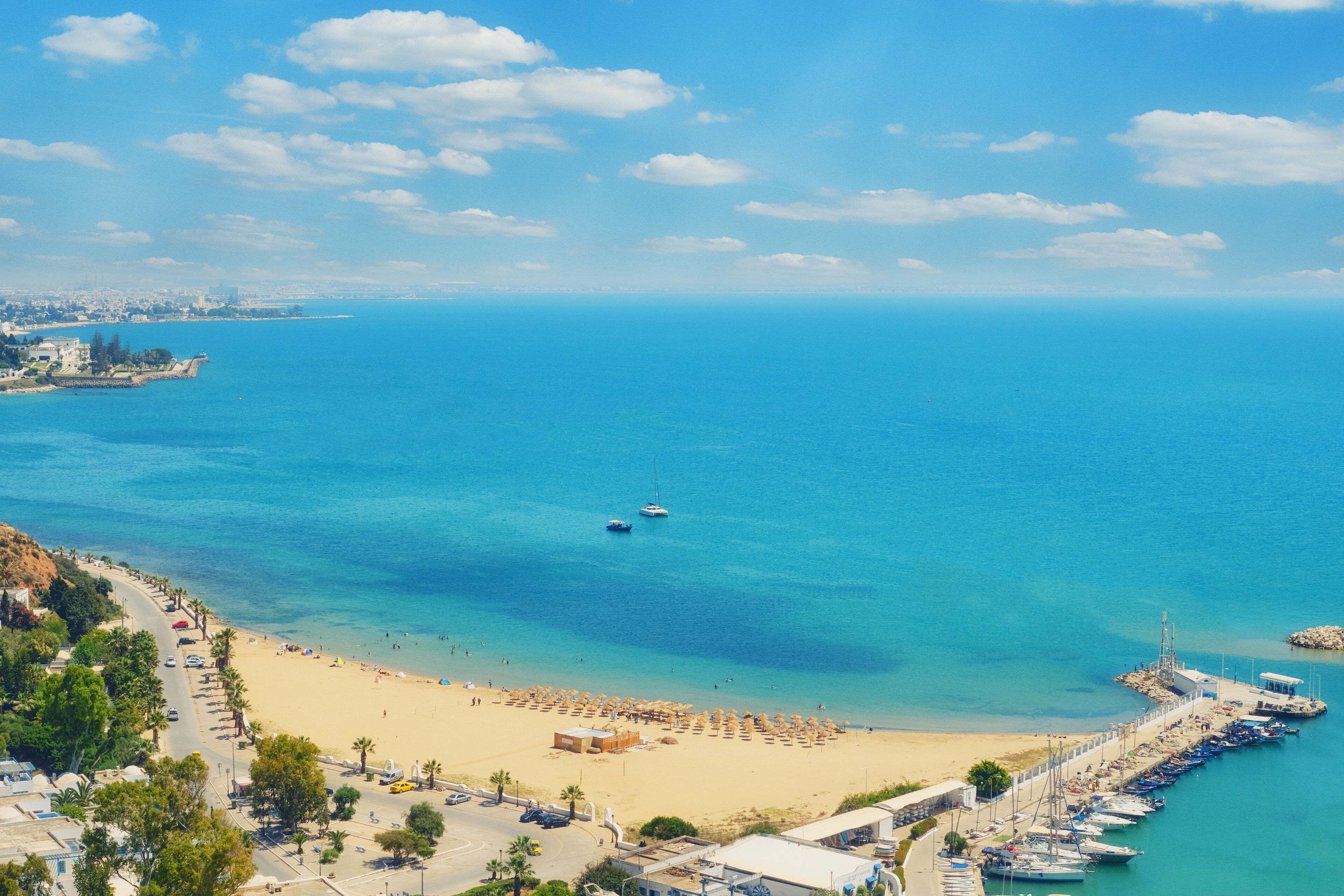 Aerial view of a sun-drenched beach with clear turquoise waters, boats moored in a small harbor, and a sweeping view of the coastline and cityscape in the distance.