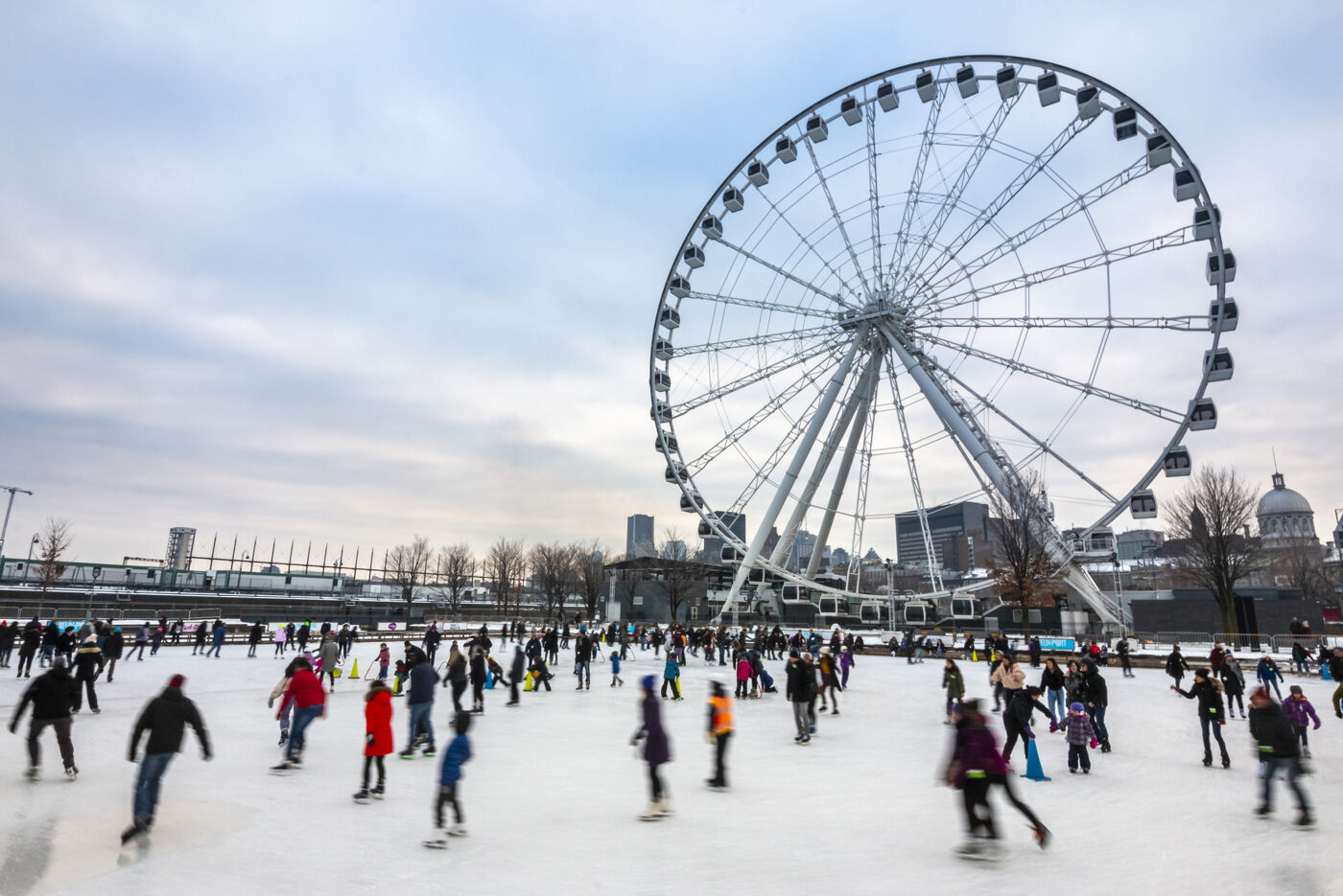 The image shows a large group of people, including children, gathered on a snowy surface, likely a skating rink or a frozen pond. They are enjoying themselves, skating and playing together in the snow.