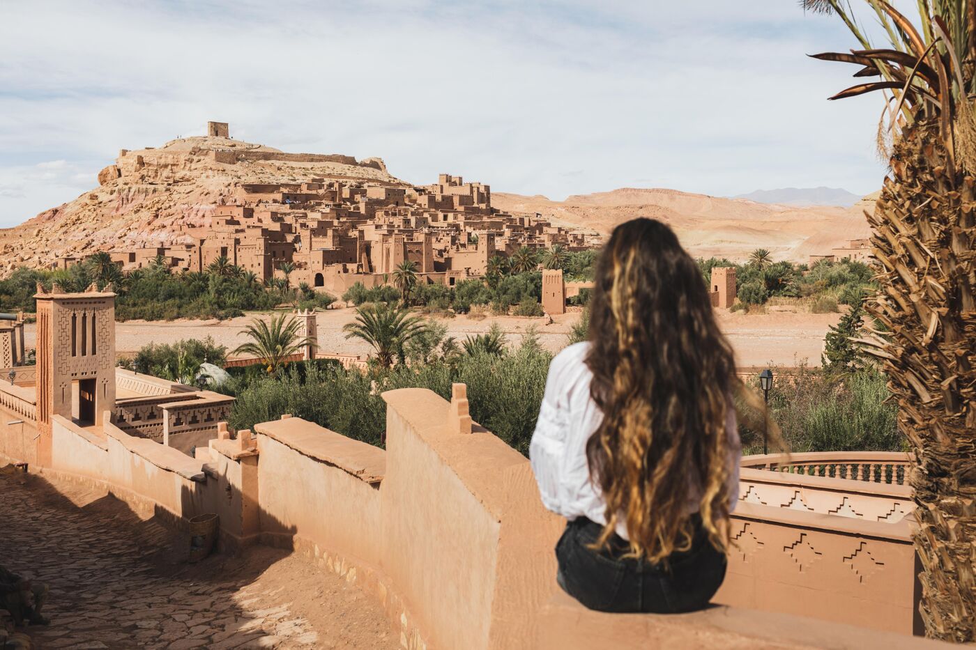 Woman sitting on fence with view of famous moroccan old town ksar Ait-Ben-Haddou. View from behind. Welcome to Morocco, Ouarzazate.