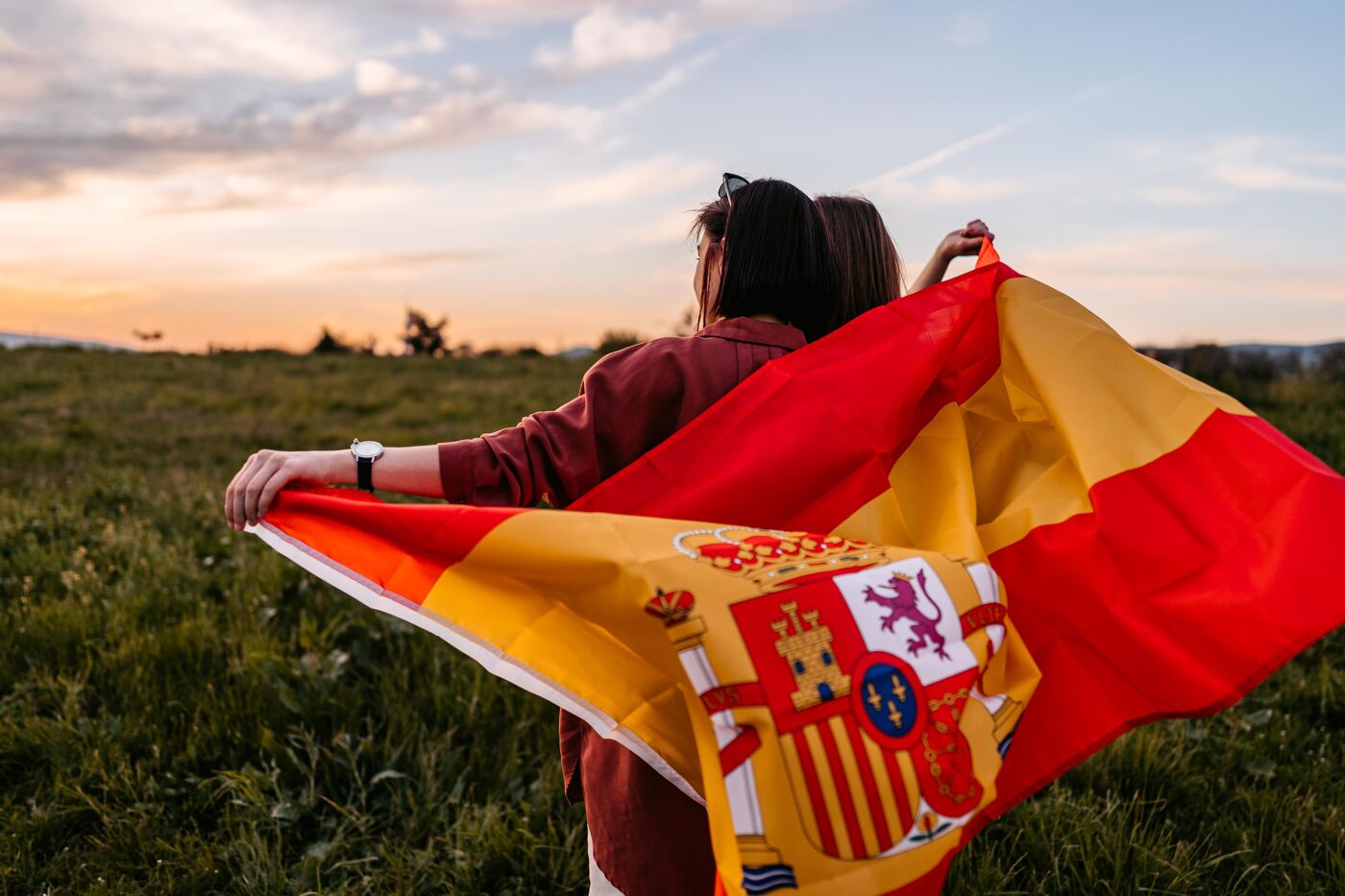 Two, young, females, covering themselves with Spanish flag. Standing on the meadow at sunset.