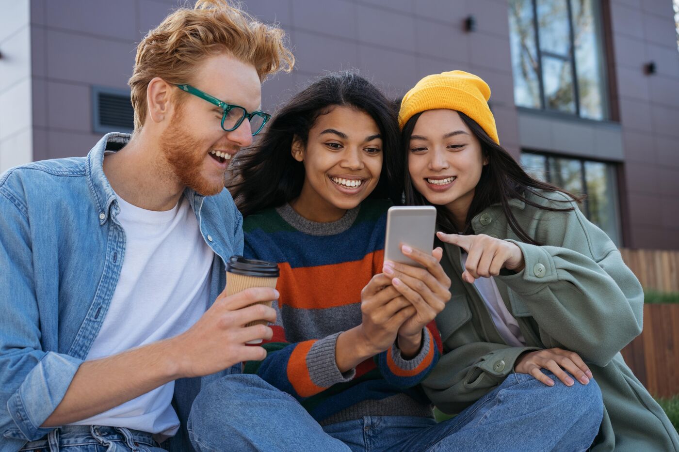 In the picture, there are three people sitting on a bench, with two of them holding a cell phone. They are smiling and enjoying their time together.