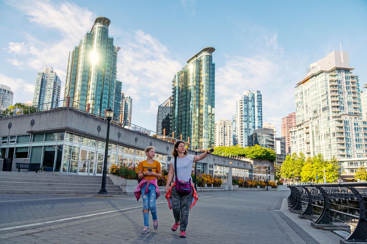 The image features a woman and a girl walking down a sidewalk in a city, with a large cityscape in the background. The woman is pointing towards the sky, possibly indicating something interesting or noteworthy in the skyline. The sidewalk is lined with trees, adding to the urban landscape.