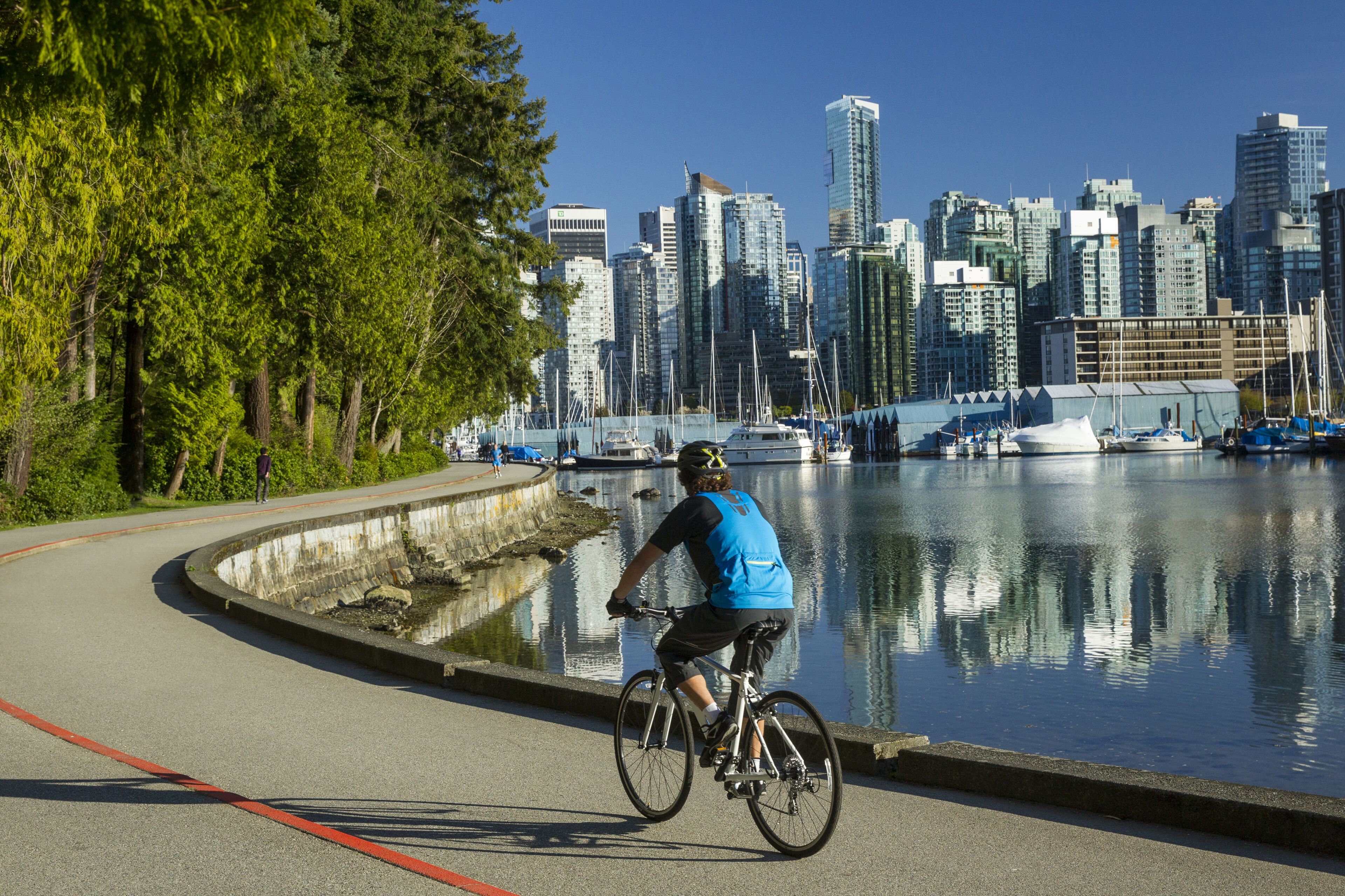 Un hombre pedalea en una bicicleta de alquiler en un día soleado de cielo azul cálido a lo largo del carril bici de la pared del mar en Stanley Park, en el centro de Vancouver, Columbia Británica, Canadá.