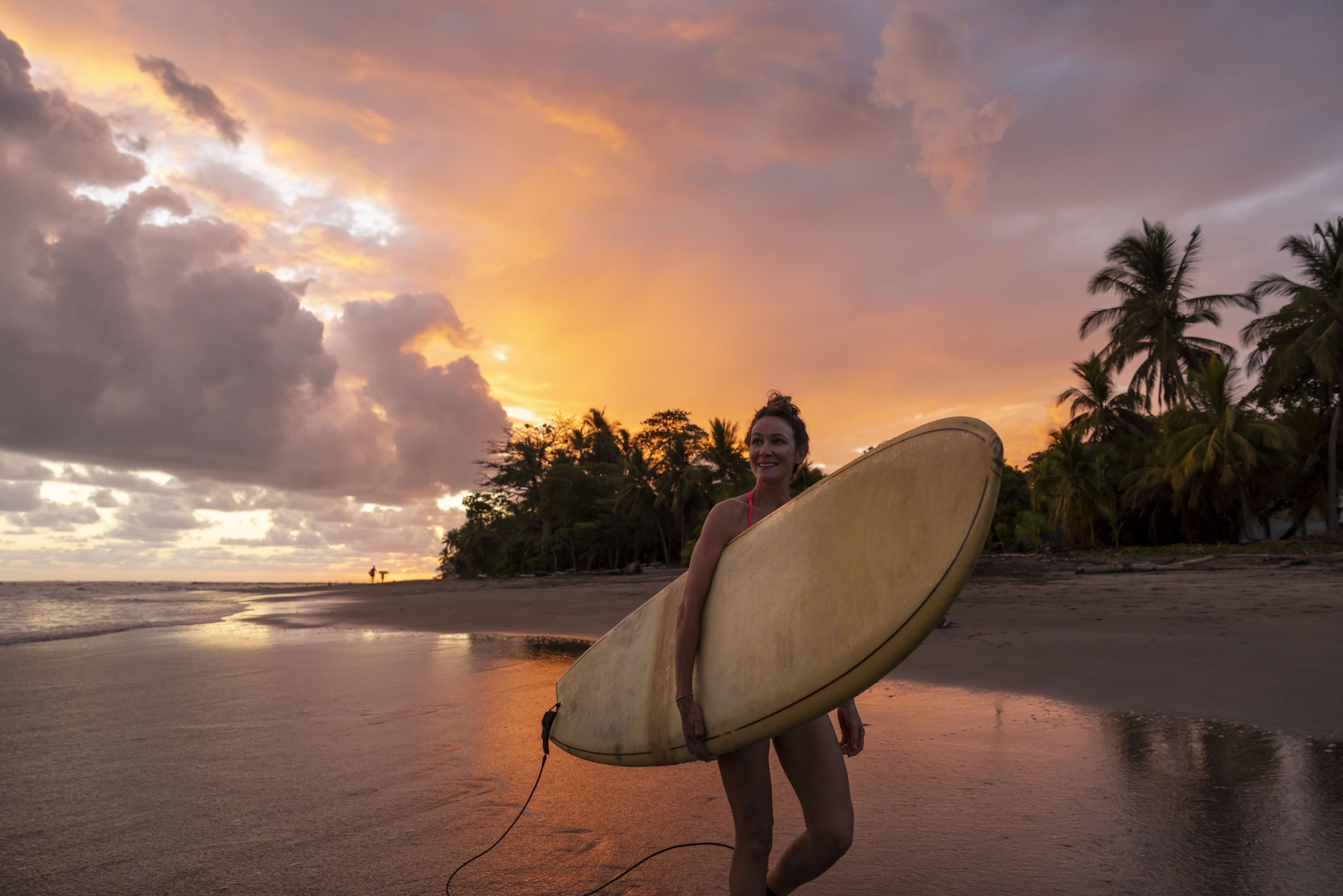 A woman walking with a surfboard during a dramatic sunset at the beach.